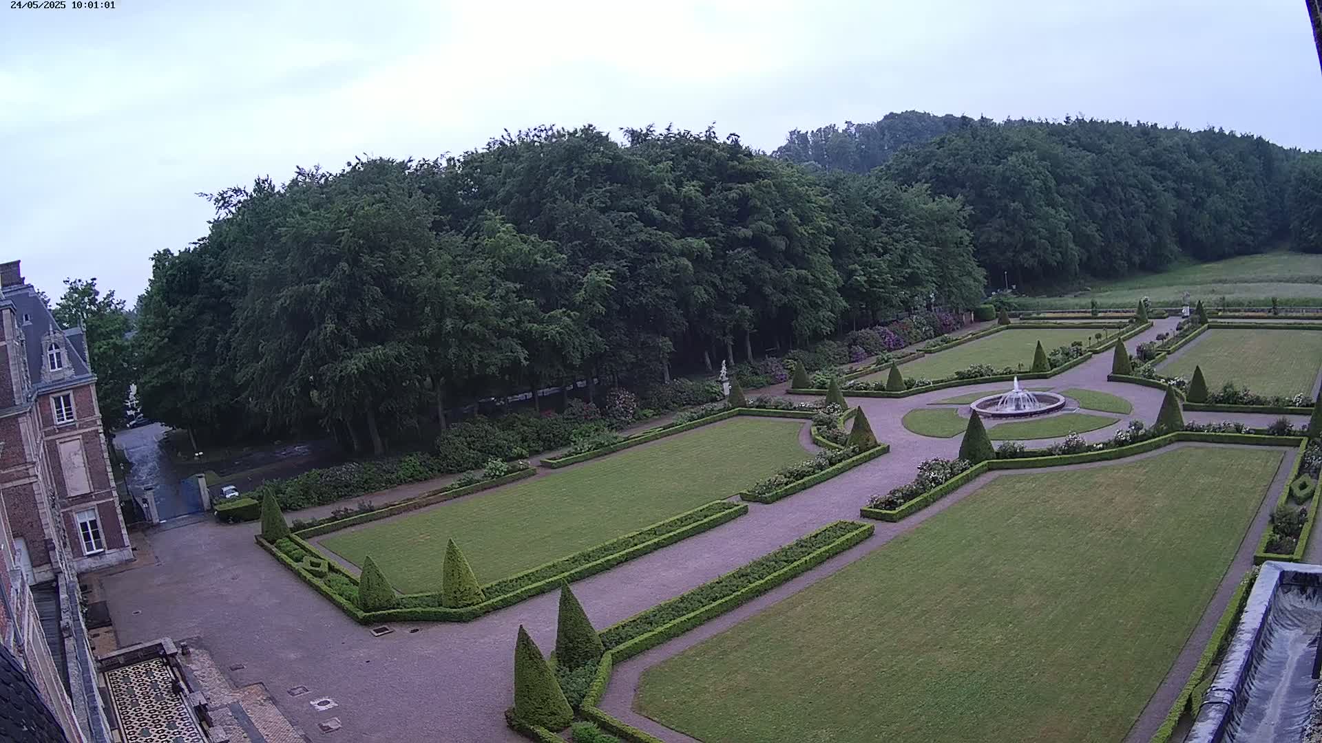 An overcast day reveals a formal garden with a fountain, manicured lawns, and hedges surrounding a chateau.