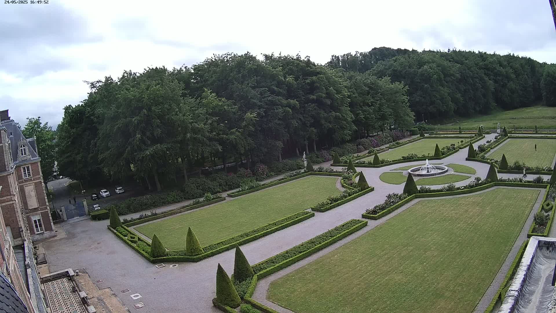 An overcast day reveals a formal garden with manicured lawns, hedges, and a central fountain, situated adjacent to a large stone building.