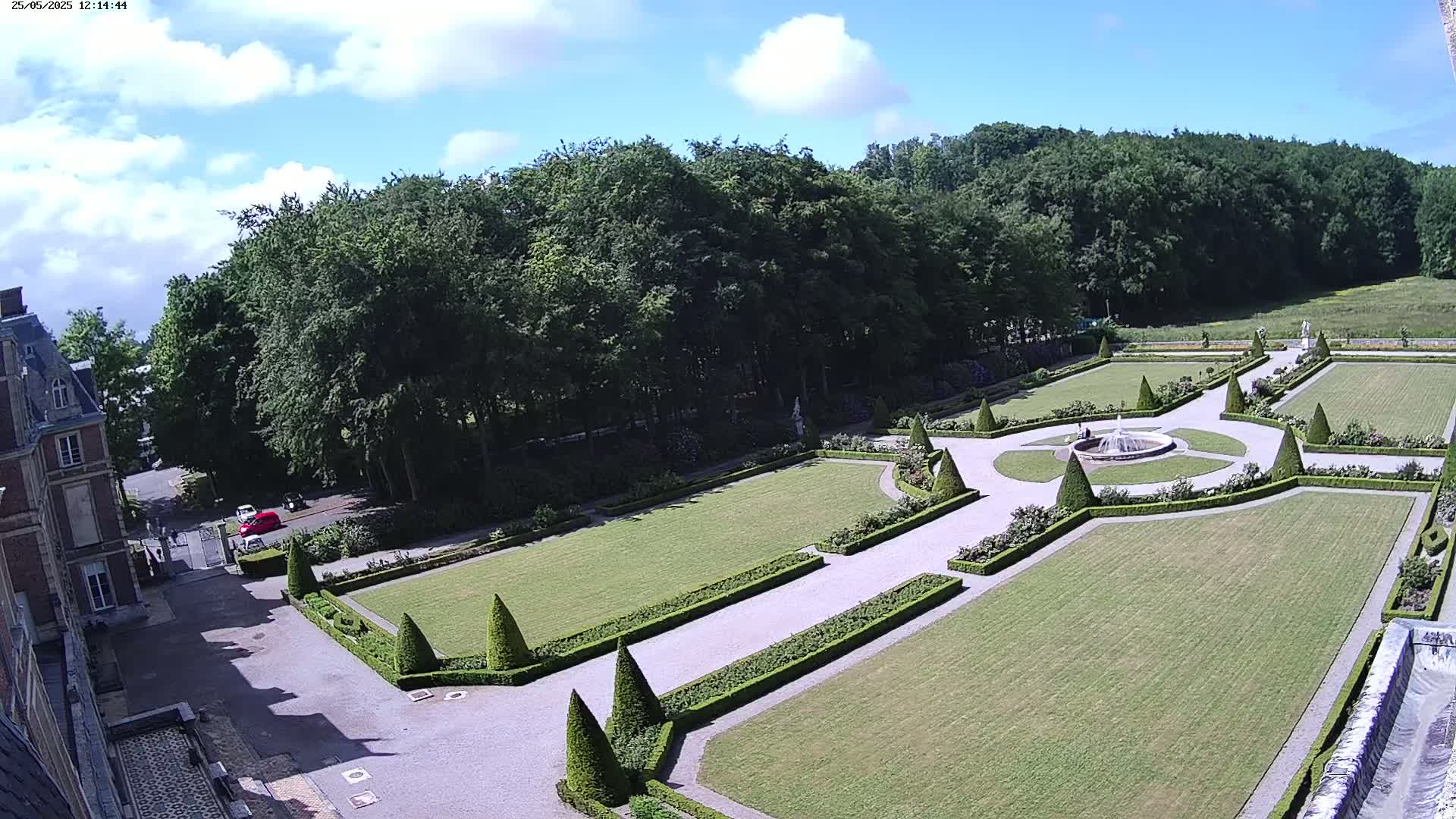 An aerial view shows a formal garden with manicured lawns, hedges, and a central fountain, bordered by a large forest under a partly cloudy sky.