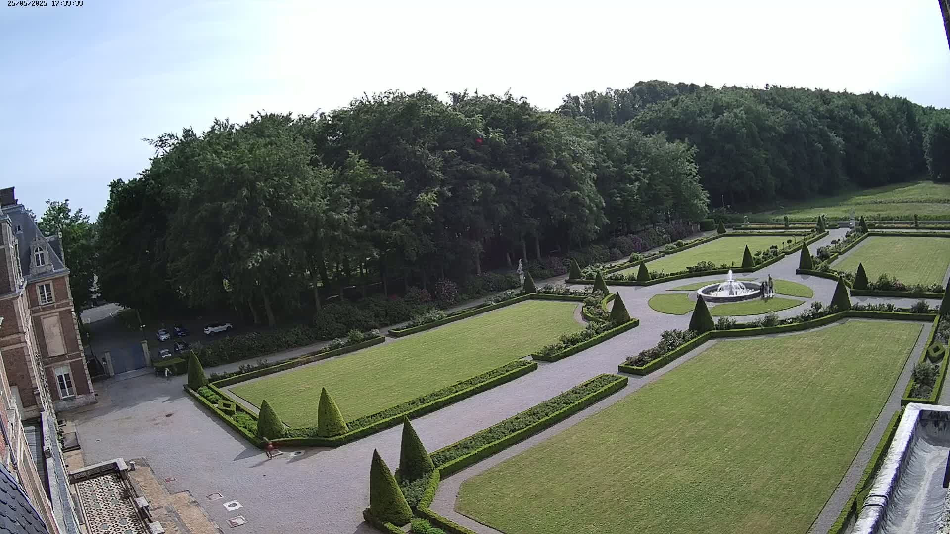 A formal garden with a fountain, manicured lawns, and hedges, is situated next to a large building under a mostly clear sky.