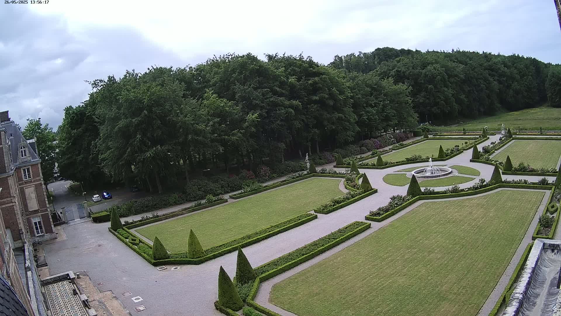 An expansive formal garden with a central fountain, meticulously manicured lawns, and neatly trimmed hedges is visible under a partly cloudy sky.