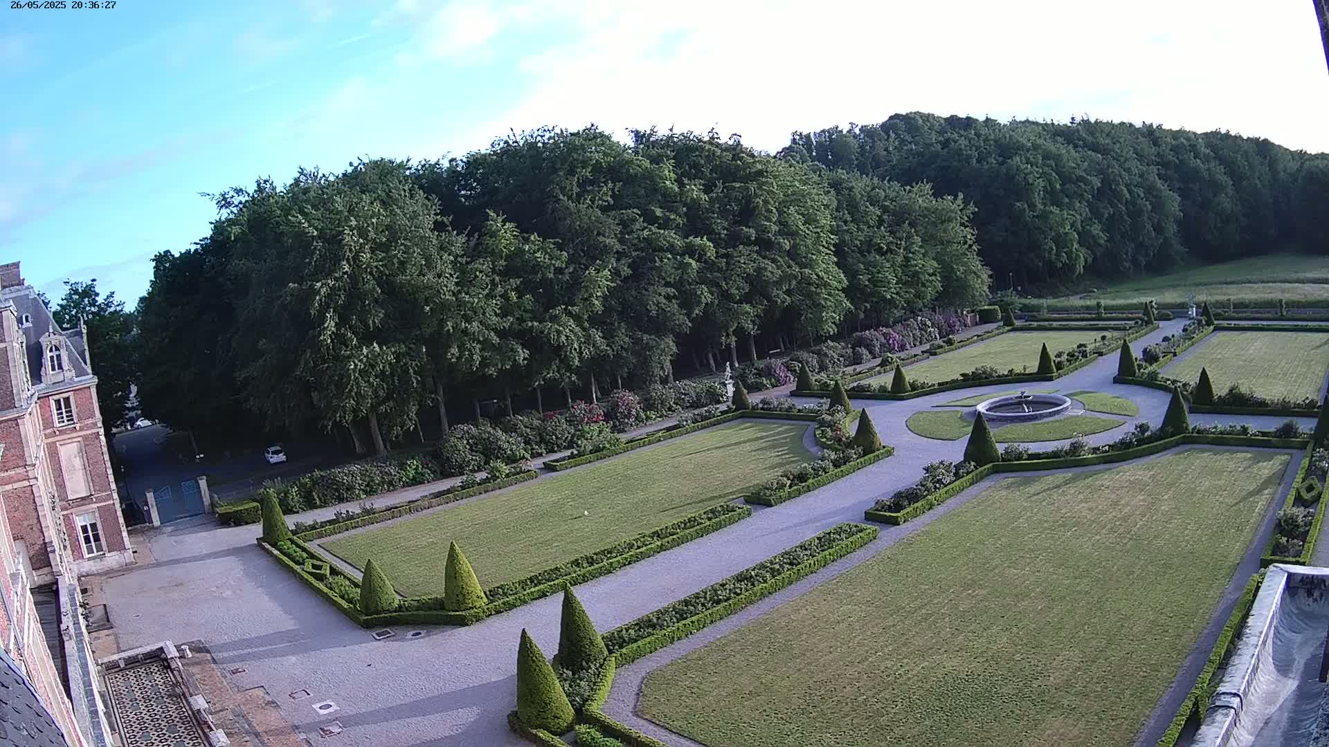 An expansive formal garden with manicured lawns, hedges, and a central fountain is situated beside a large brick building under a clear, bright sky.