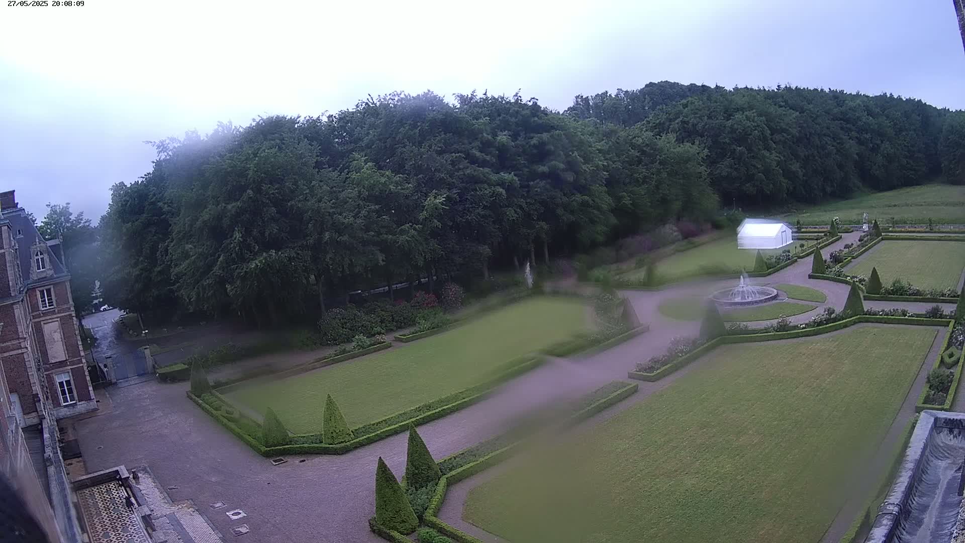 An elevated view of a formal garden with a fountain, hedges, and a white tent, situated beside a brick building under a cloudy, possibly rainy sky.