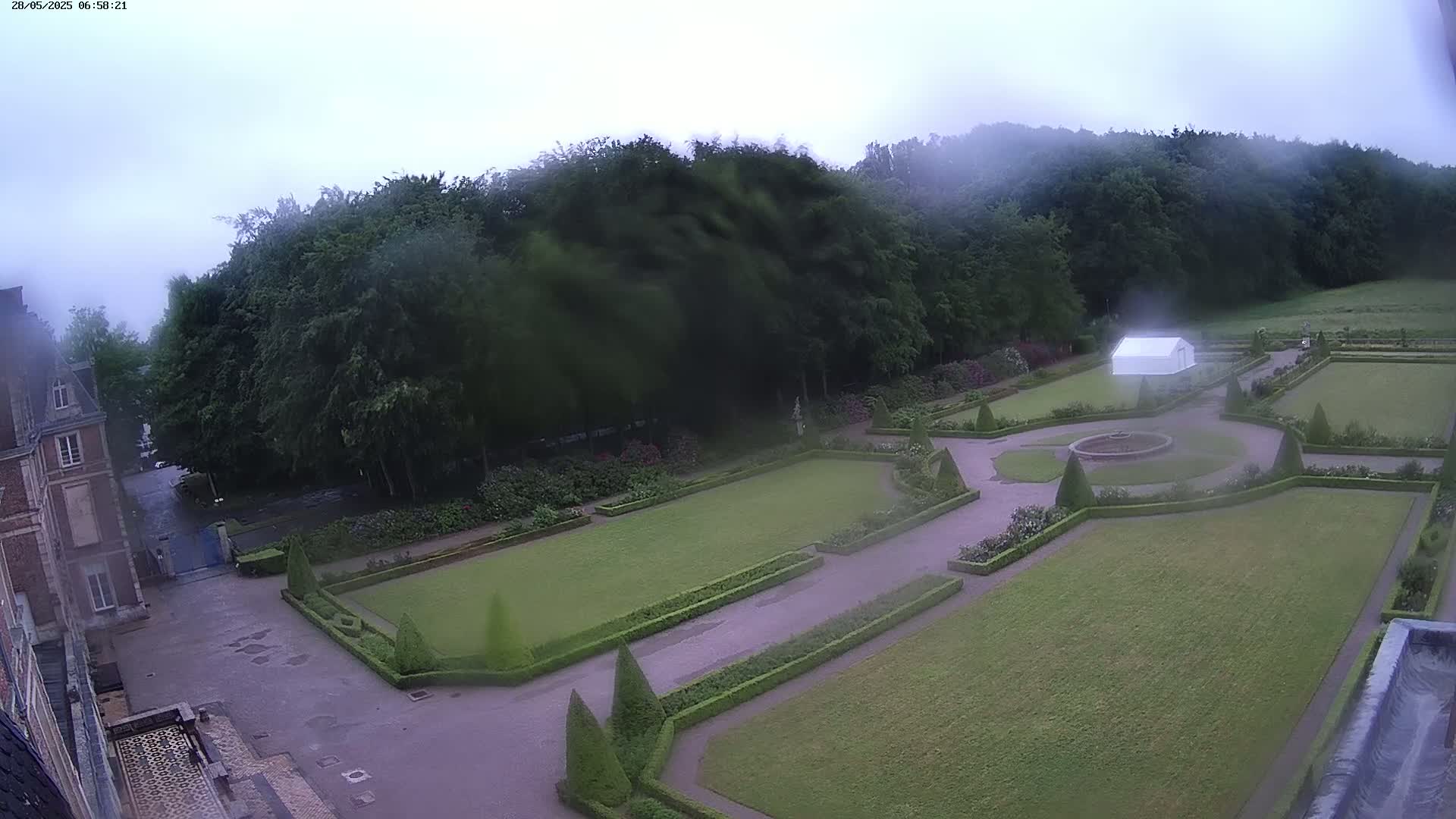 A formal garden with manicured lawns, hedges, and a fountain is visible, overlooked by a building, all under a cloudy sky.