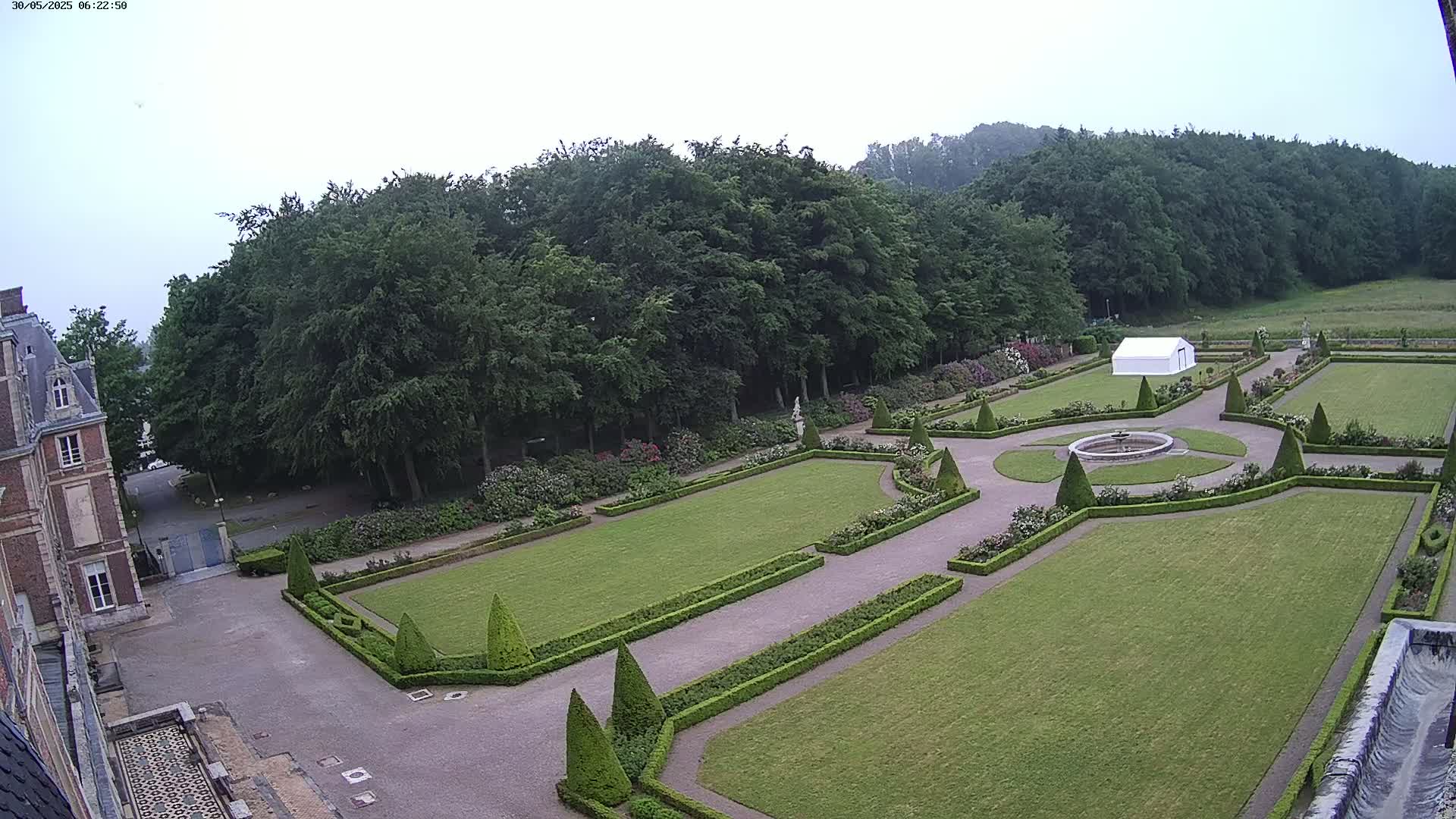 An overcast day reveals a formal garden with manicured lawns, hedges, and a central fountain, adjacent to a large brick building and a wooded area.