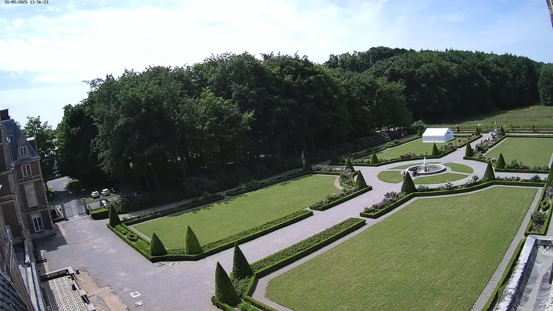 An aerial view shows a formal garden with manicured lawns, hedges, and a fountain, adjacent to a large building under a mostly clear, sunny sky.