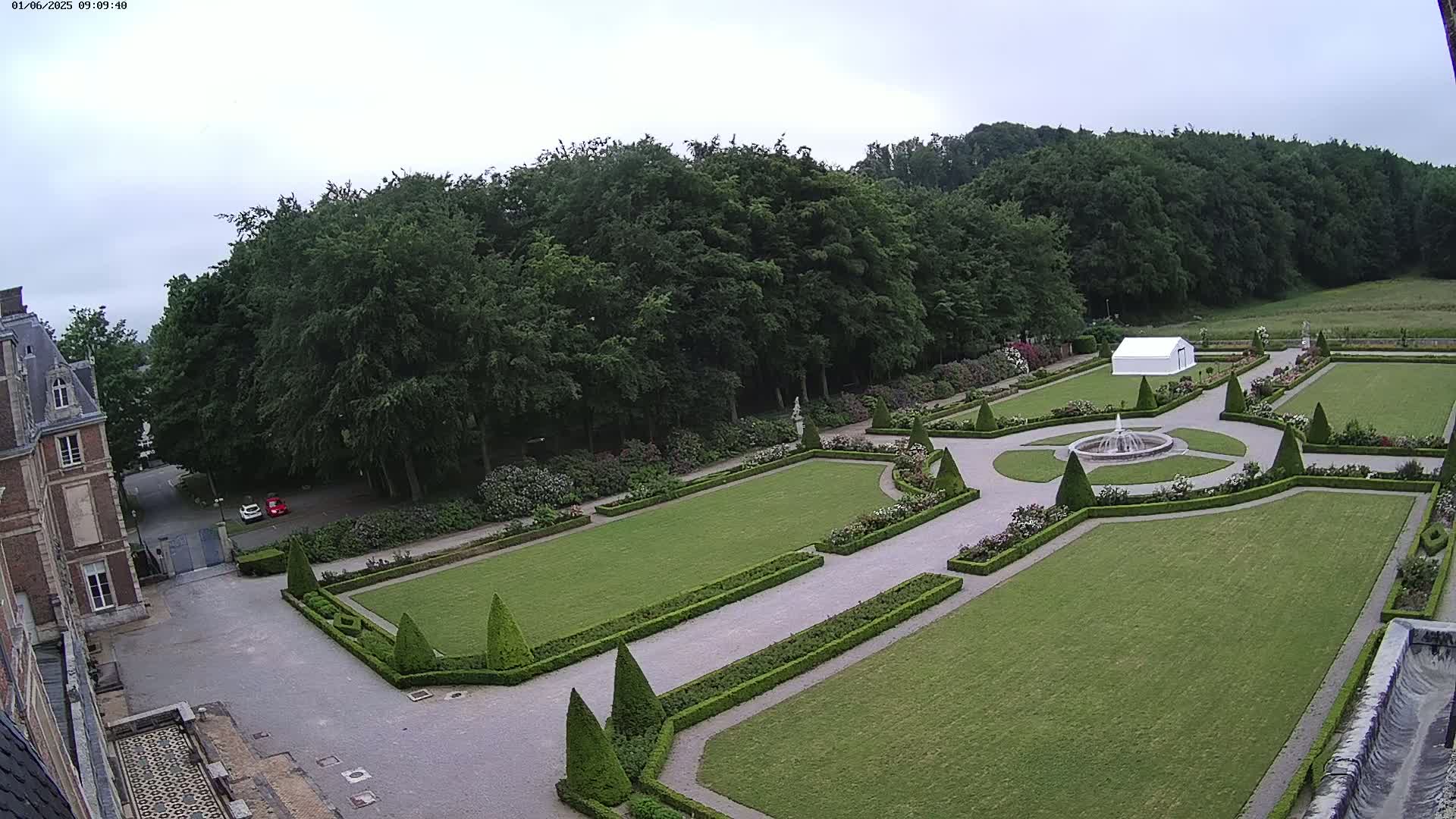 An overcast day reveals a formal garden with manicured lawns, hedges, and a fountain, situated next to a large building and bordered by a forest.