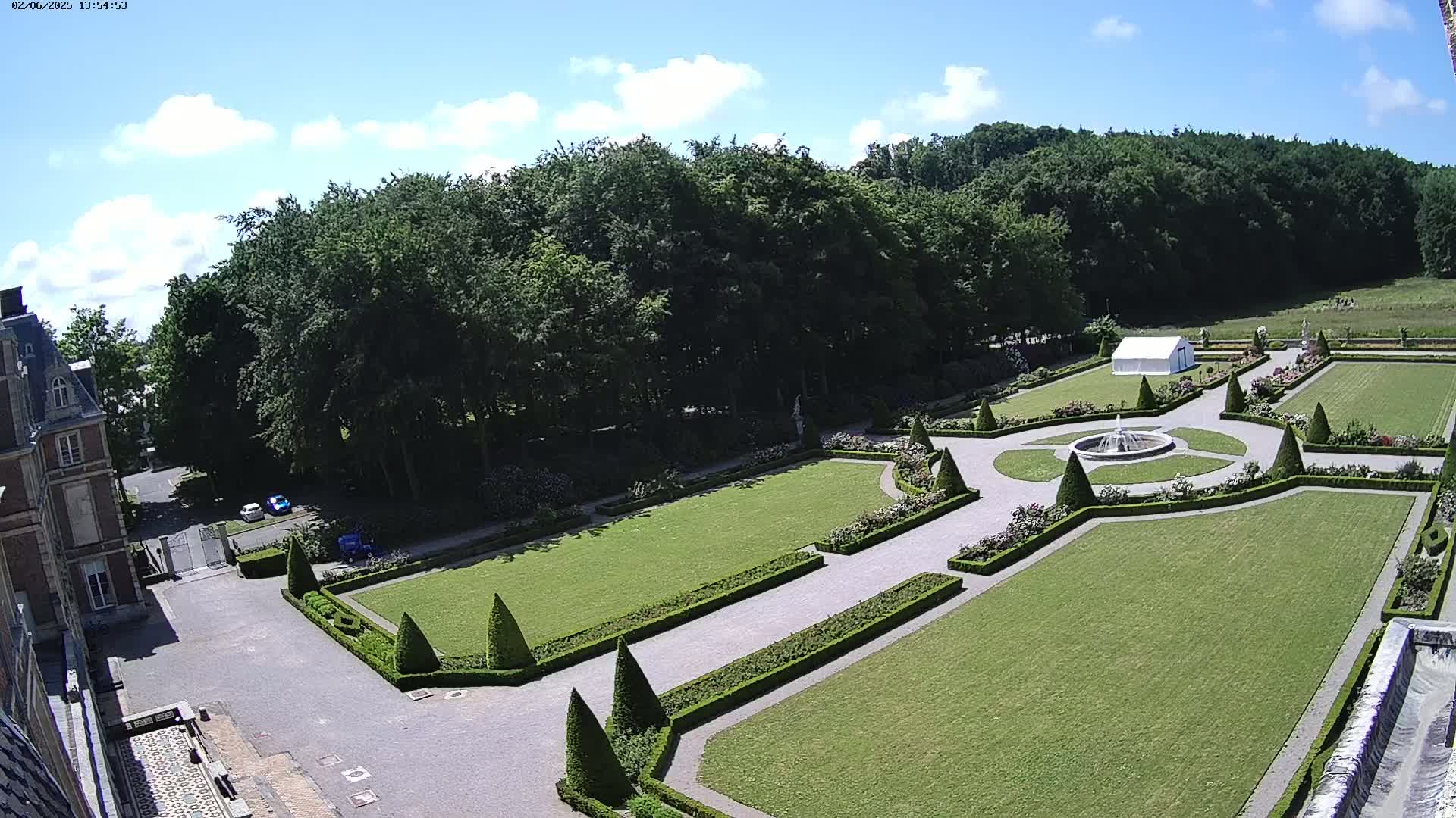 An elevated view shows a formal garden with manicured lawns, hedges, and a fountain, adjacent to a large building under a partly cloudy sky.
