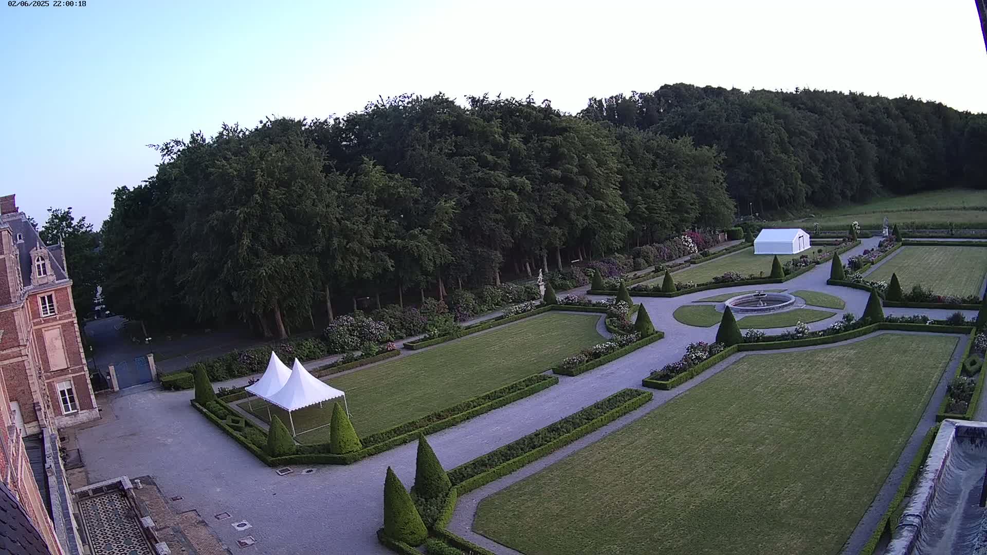 An expansive formal garden with manicured lawns, hedges, and a central fountain, situated next to a large brick building under a twilight sky.