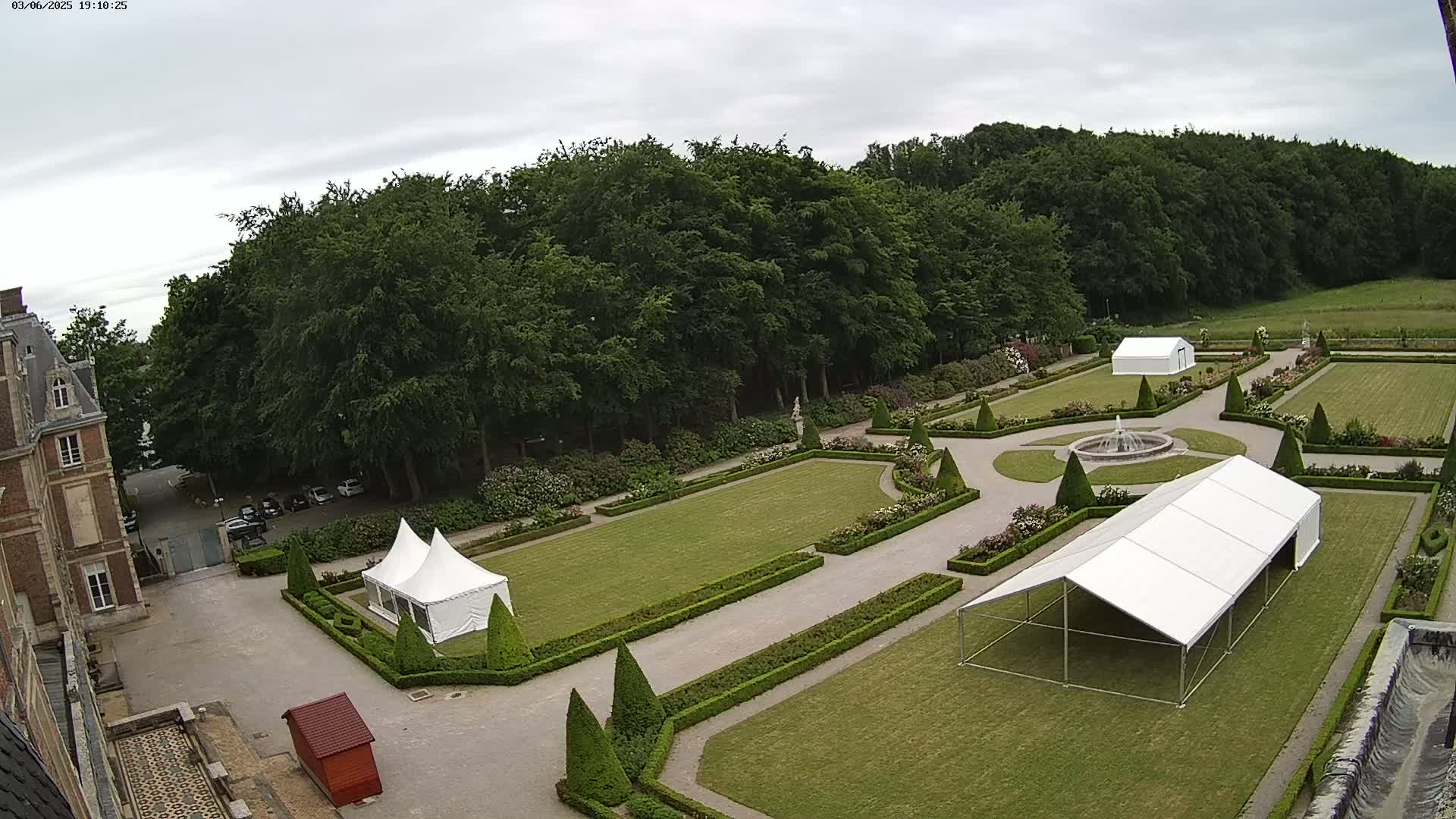 An overcast day reveals a formal garden with manicured lawns, hedges, and flowerbeds surrounding a fountain, and two white tents situated near a large building.