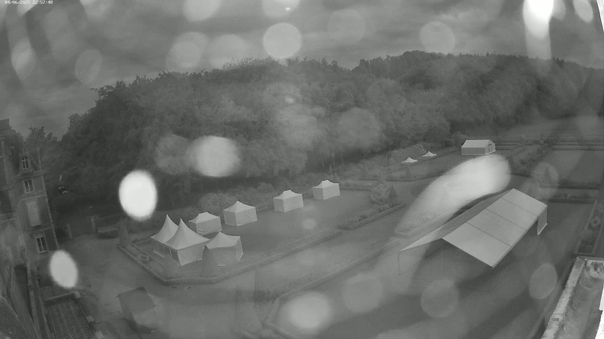 Several white tents are set up in a rain-soaked formal garden next to a large building, with trees visible in the background.