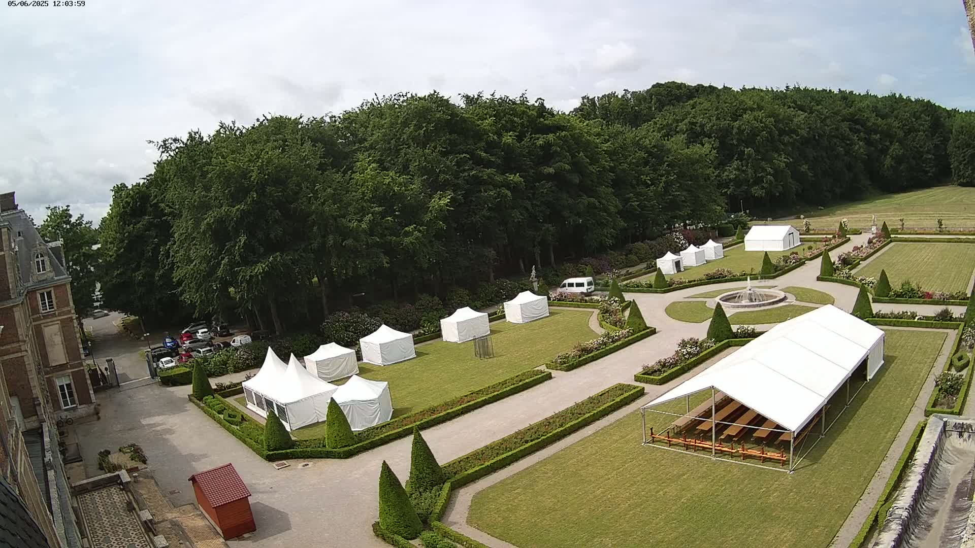 A partly cloudy day reveals a formal garden with several white tents set up on the manicured lawn, near a chateau and surrounding woods.