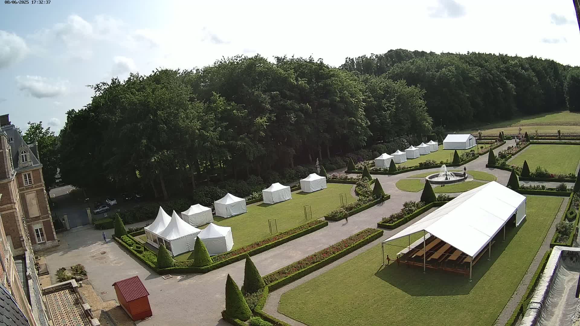 An aerial view shows numerous white tents set up on a manicured lawn adjacent to a chateau on a partly cloudy day.