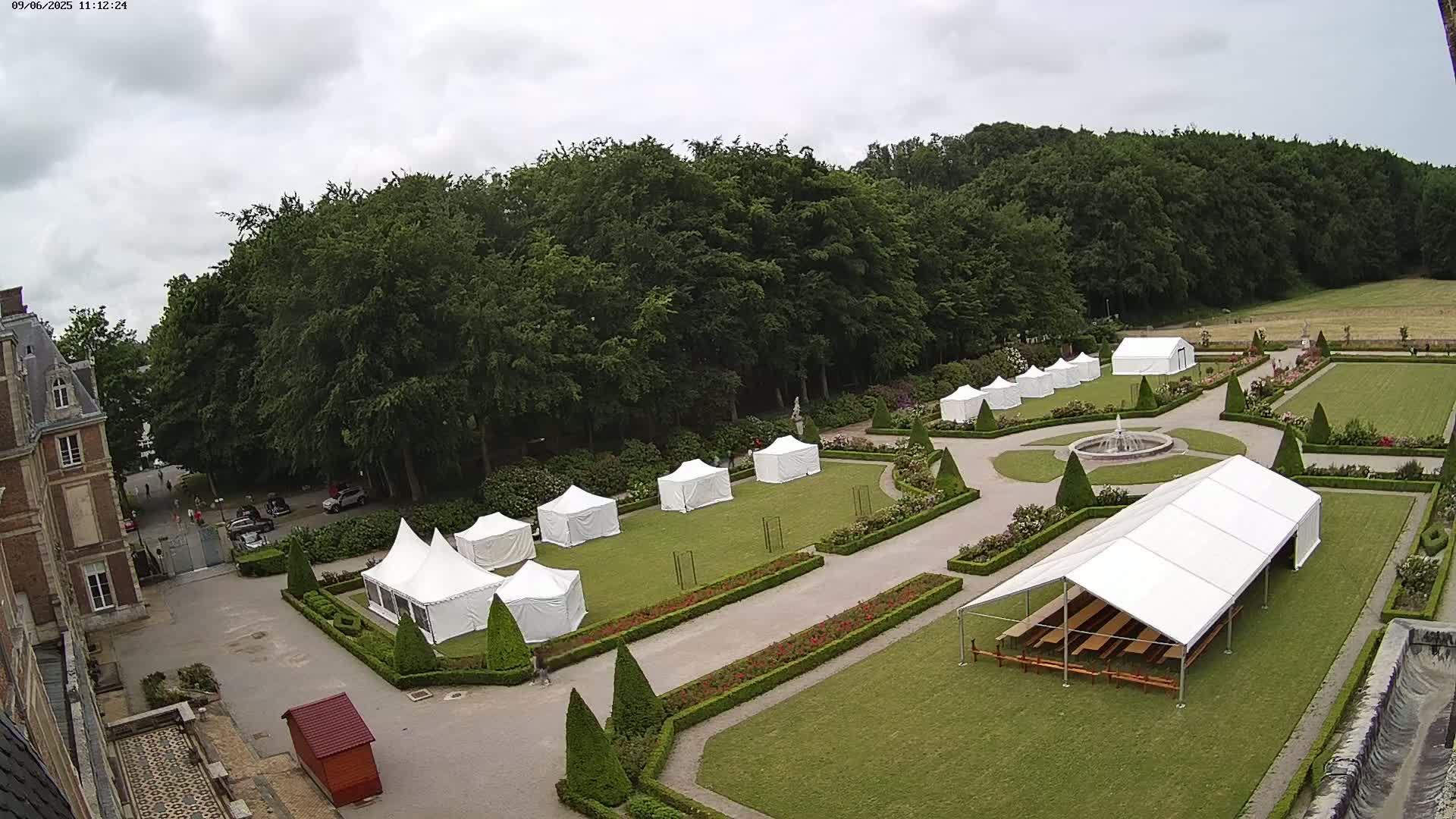 A large, formal garden with several white tents set up on the lawn, under a mostly cloudy sky, next to a chateau.