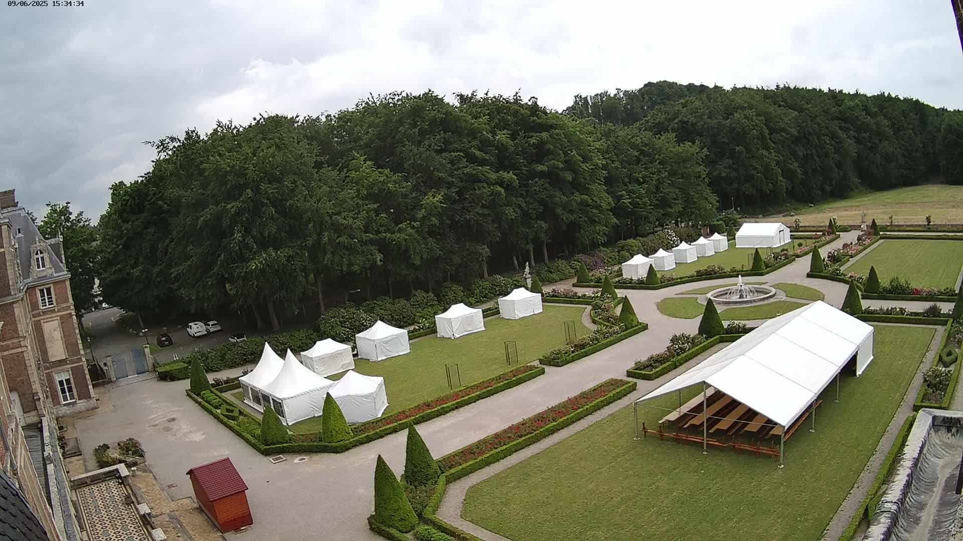 An overcast day shows numerous white tents set up on a manicured lawn, surrounding a fountain, next to a large building.