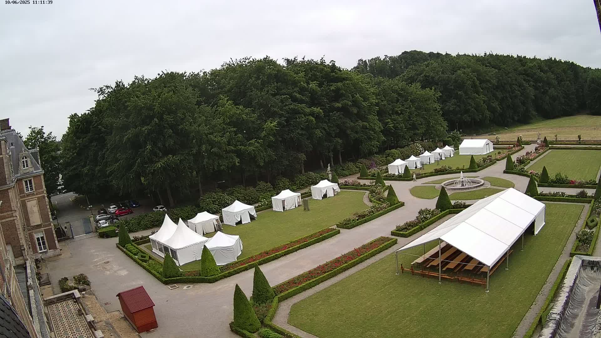 An overcast day shows a large, formal garden with numerous white tents set up on the lawn near a chateau.