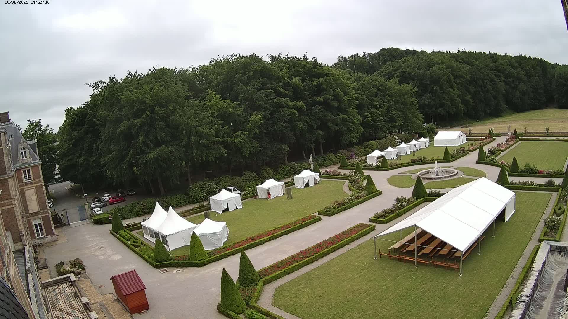 A high-angle view shows a chateau's formal gardens on an overcast day with numerous white tents set up on the manicured lawns.