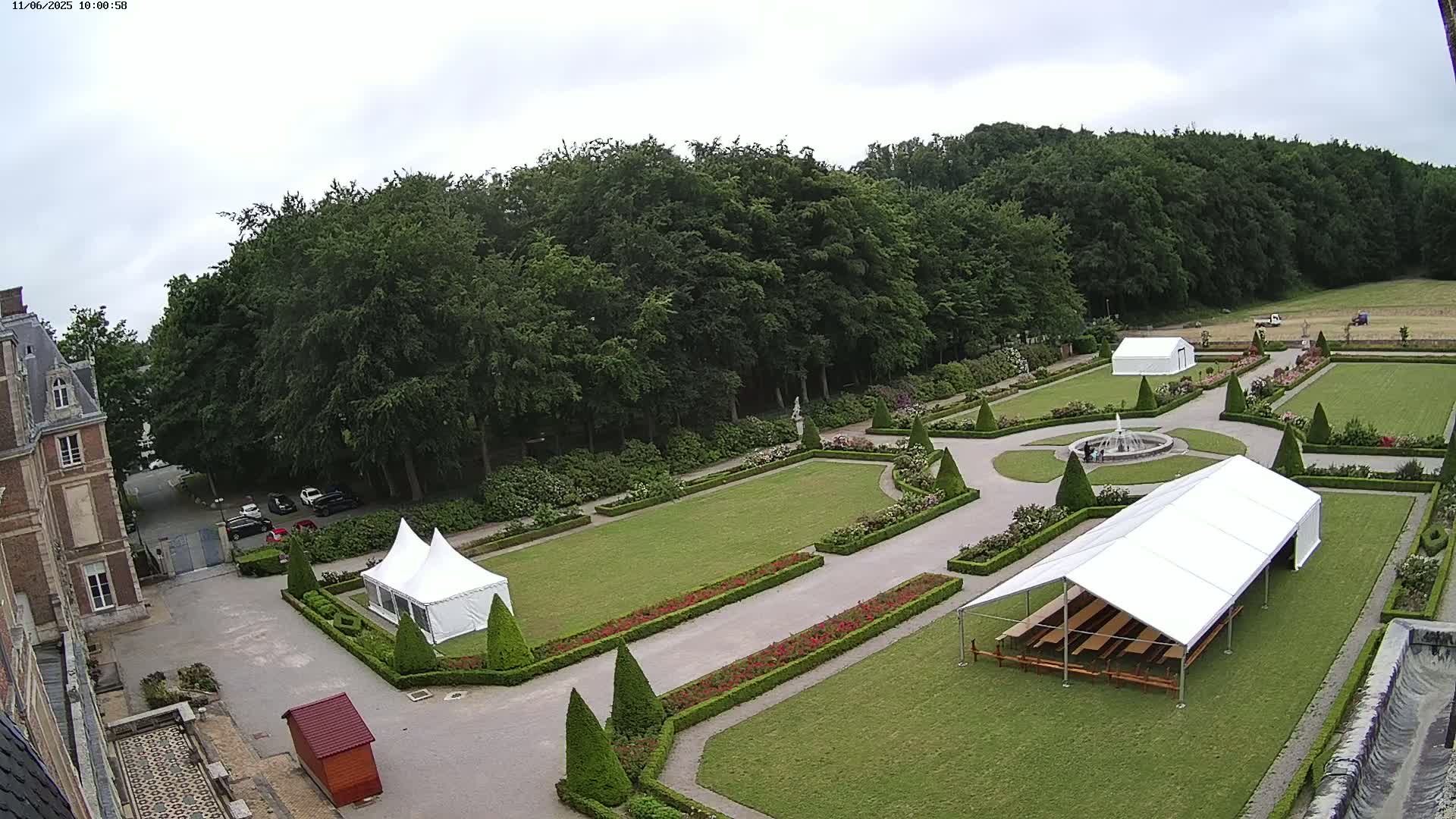 An aerial view shows a formal garden with manicured lawns, hedges, and two white tents near a chateau on an overcast day.