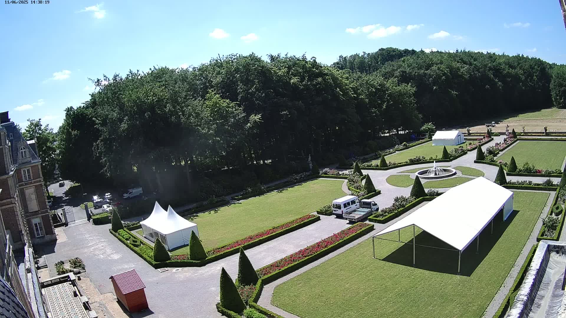 An aerial view shows a formal garden with two white tents, a fountain, manicured lawns, and hedges, all situated next to a large building under a sunny sky.