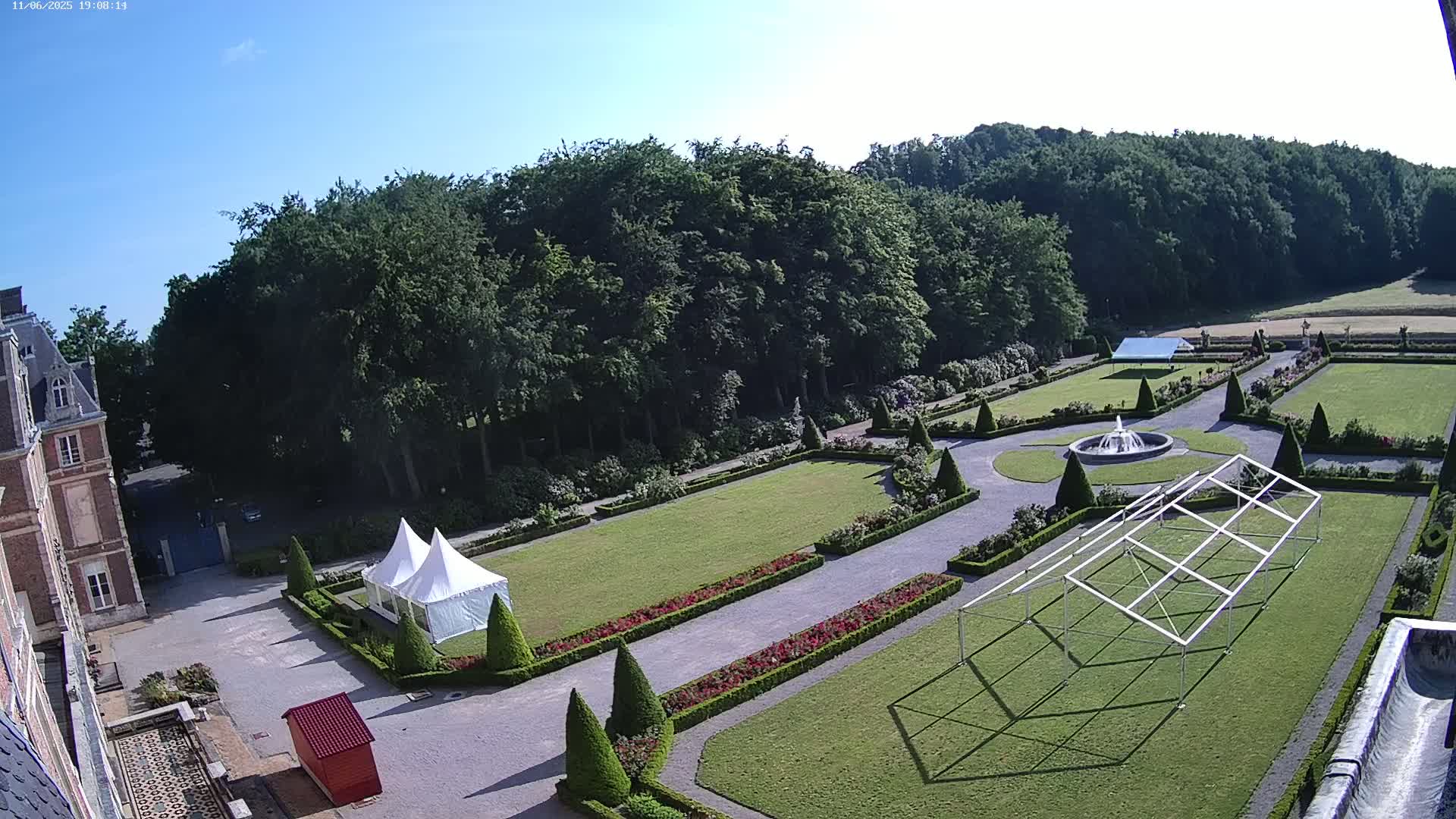 An expansive formal garden, under a clear blue sky, features manicured lawns, flowerbeds, a fountain, and two party tents near a large brick building.