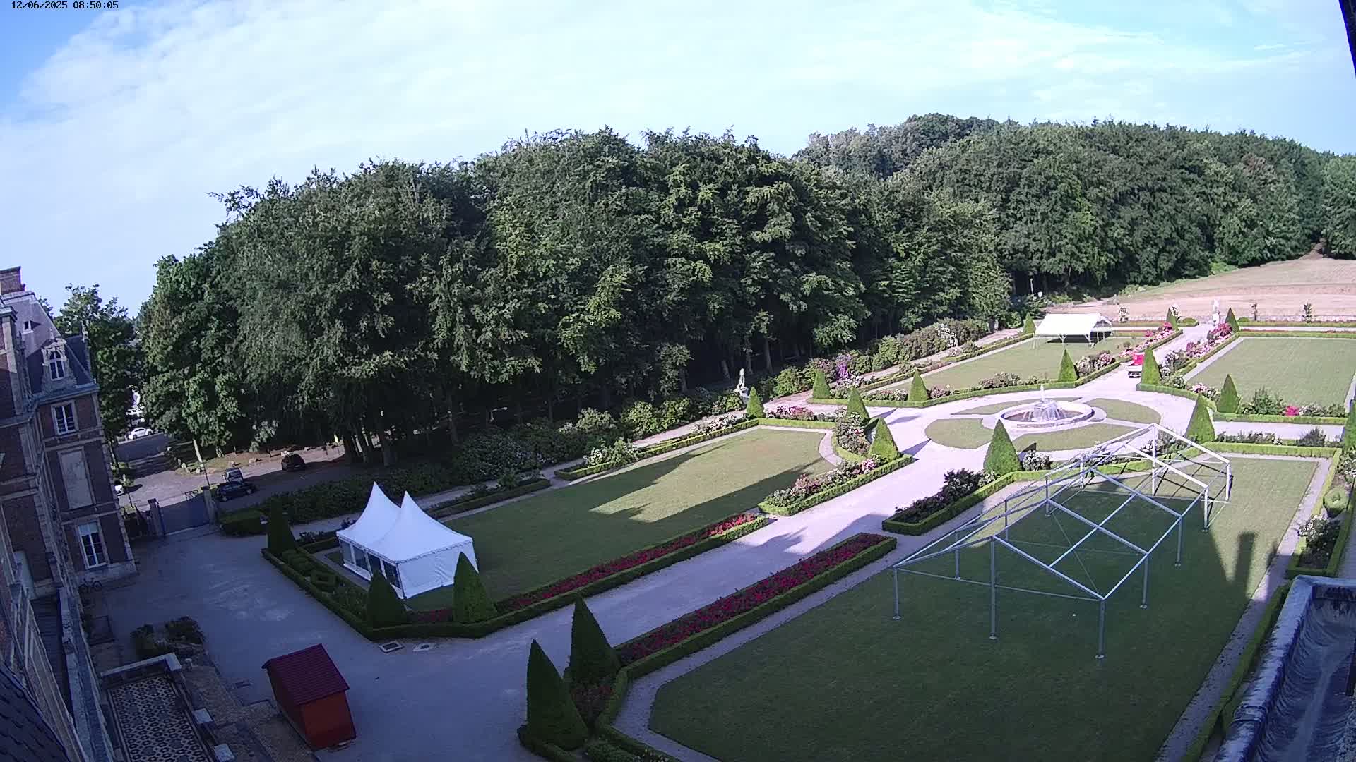 A large formal garden with manicured lawns, hedges, and flowerbeds surrounds a chateau on a partly sunny day.