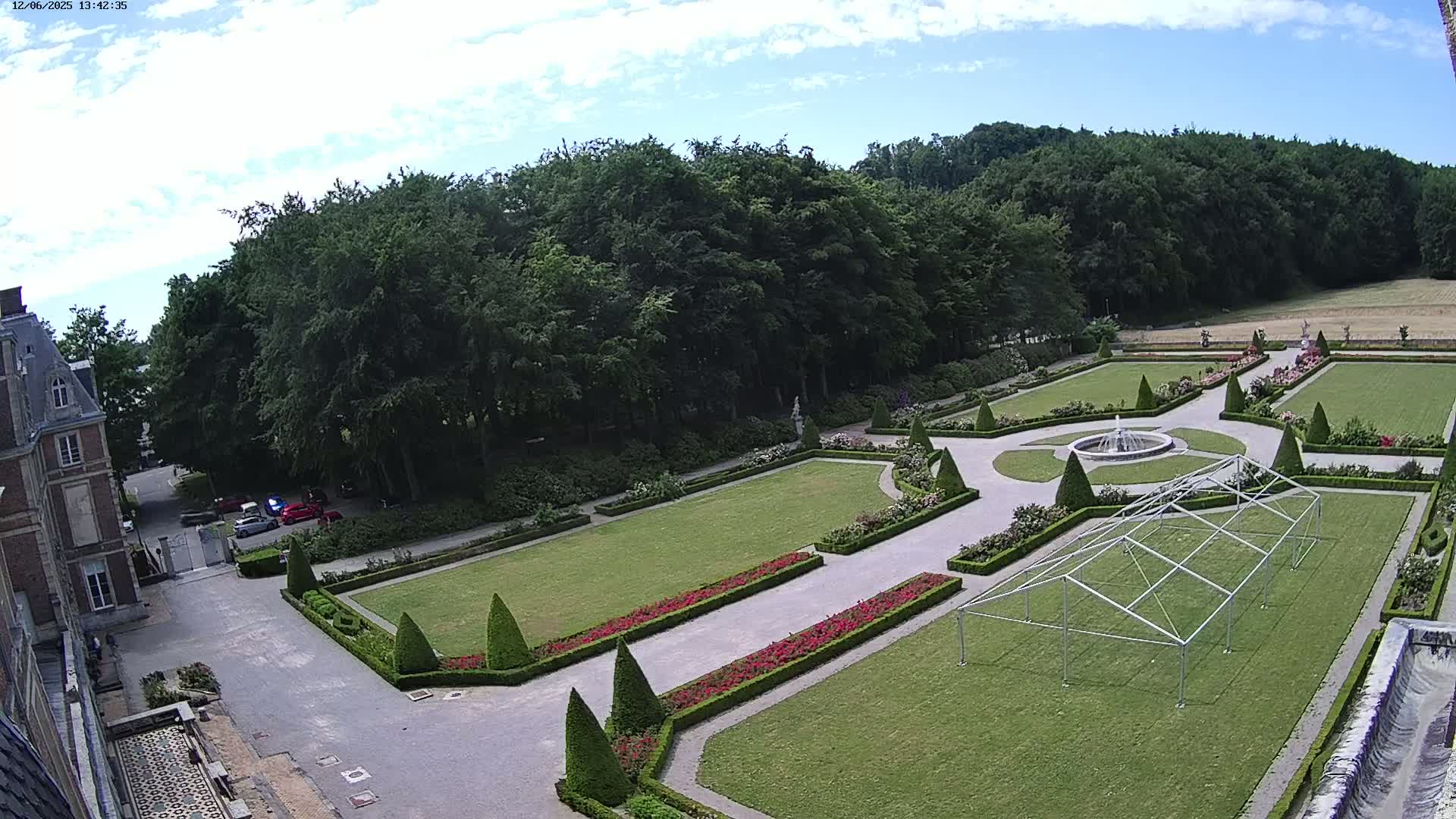 An aerial view shows a formal garden with manicured lawns, hedges, and a fountain, adjacent to a large building under a mostly sunny sky.