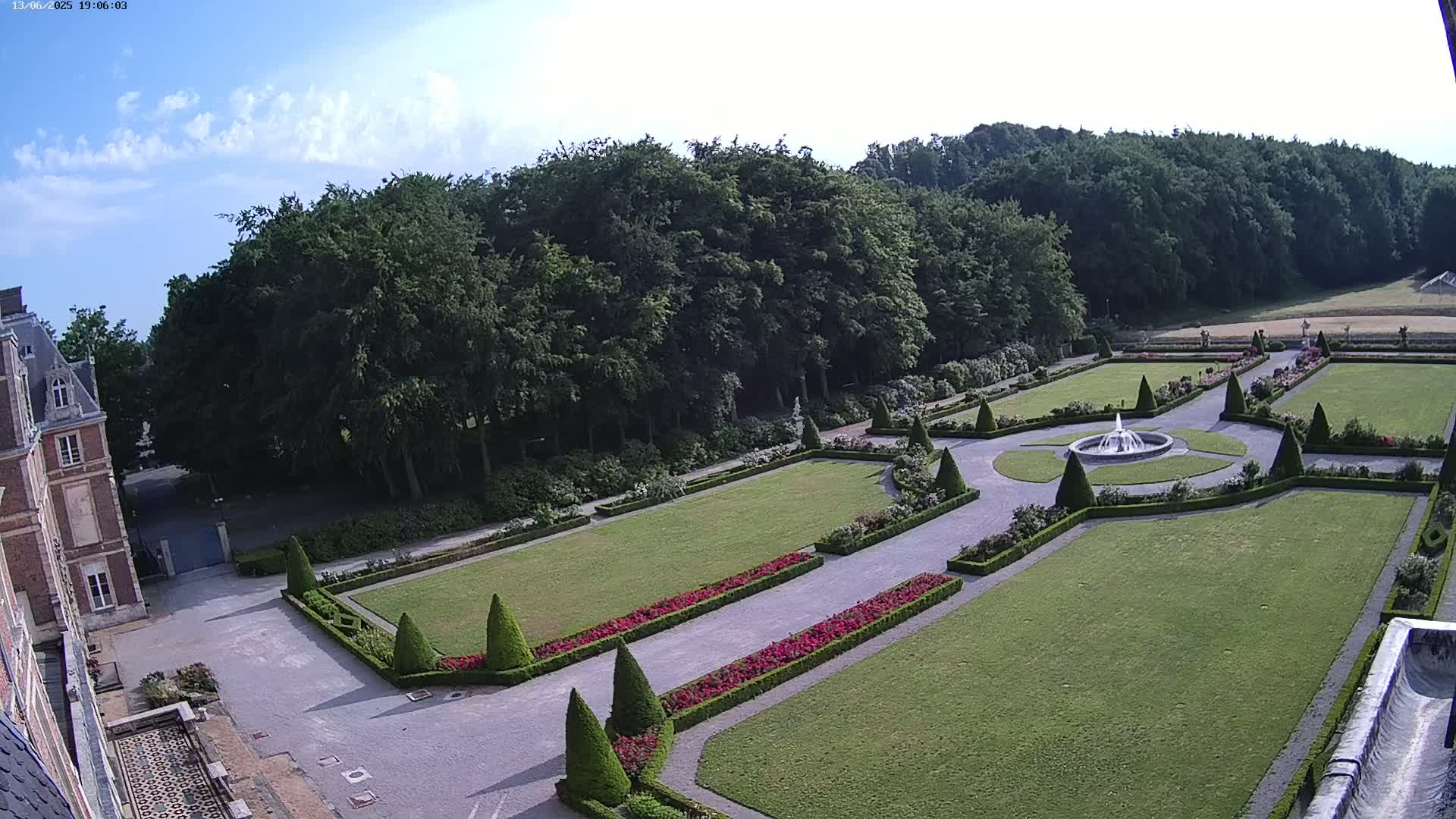 An aerial view shows a formal garden with a fountain, manicured lawns, and hedges, adjacent to a large brick building under a partly cloudy sky.