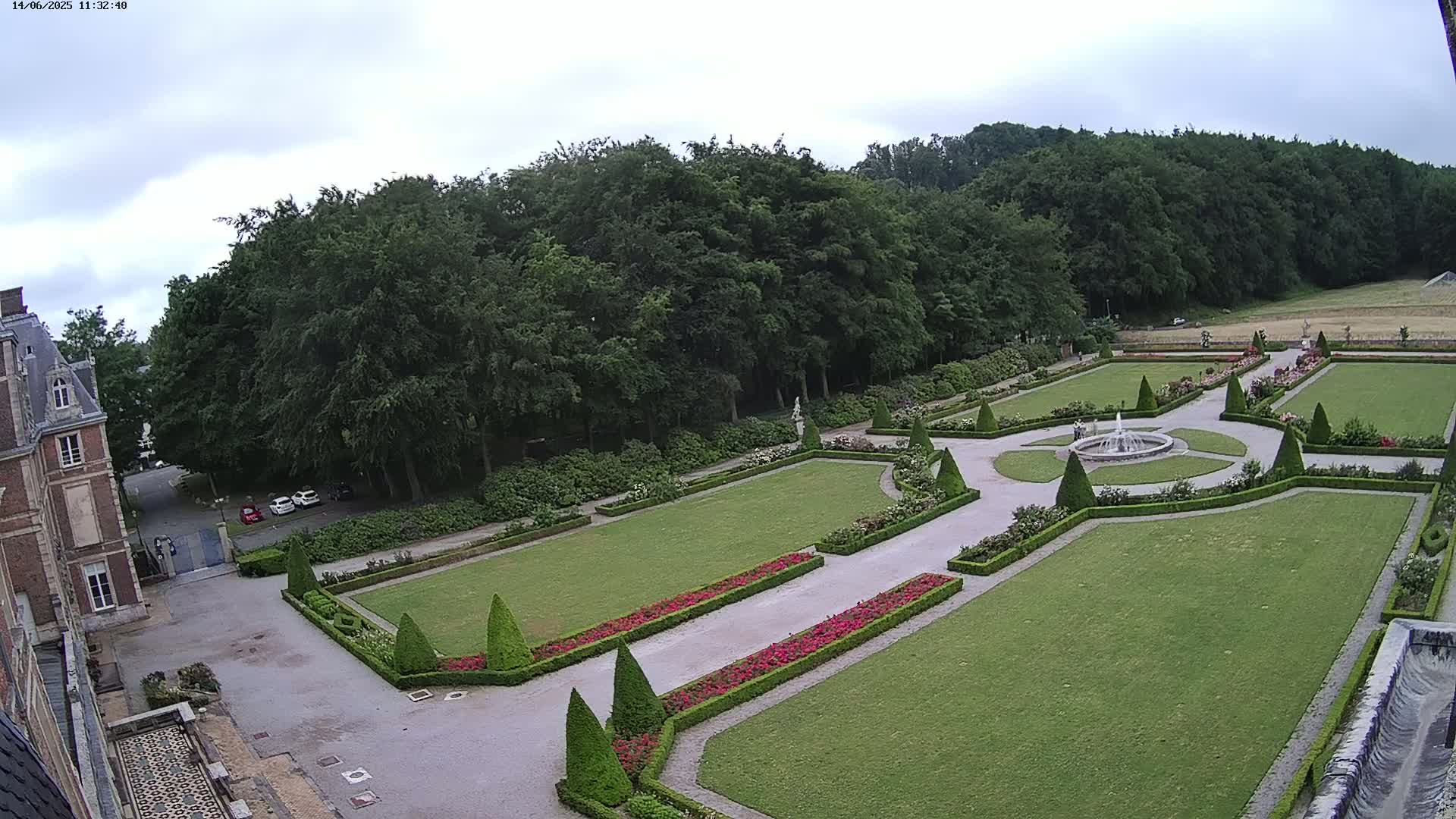 An expansive formal garden with a fountain, manicured lawns, and hedges, bordered by a large building and a forest under an overcast sky.