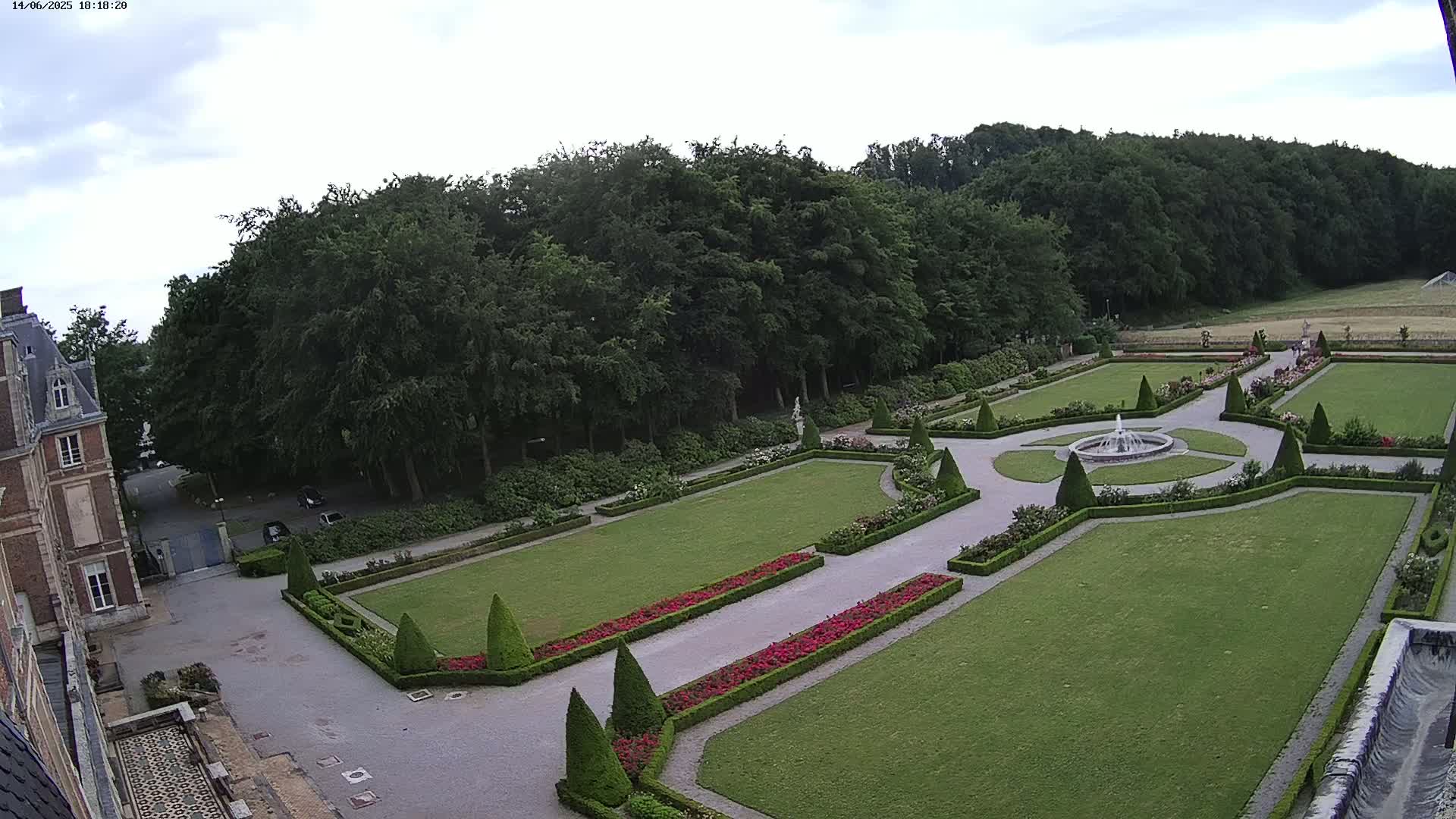 An aerial view shows an expansive formal garden with a central fountain, manicured lawns, and hedges, adjacent to a large building under a partly cloudy sky.
