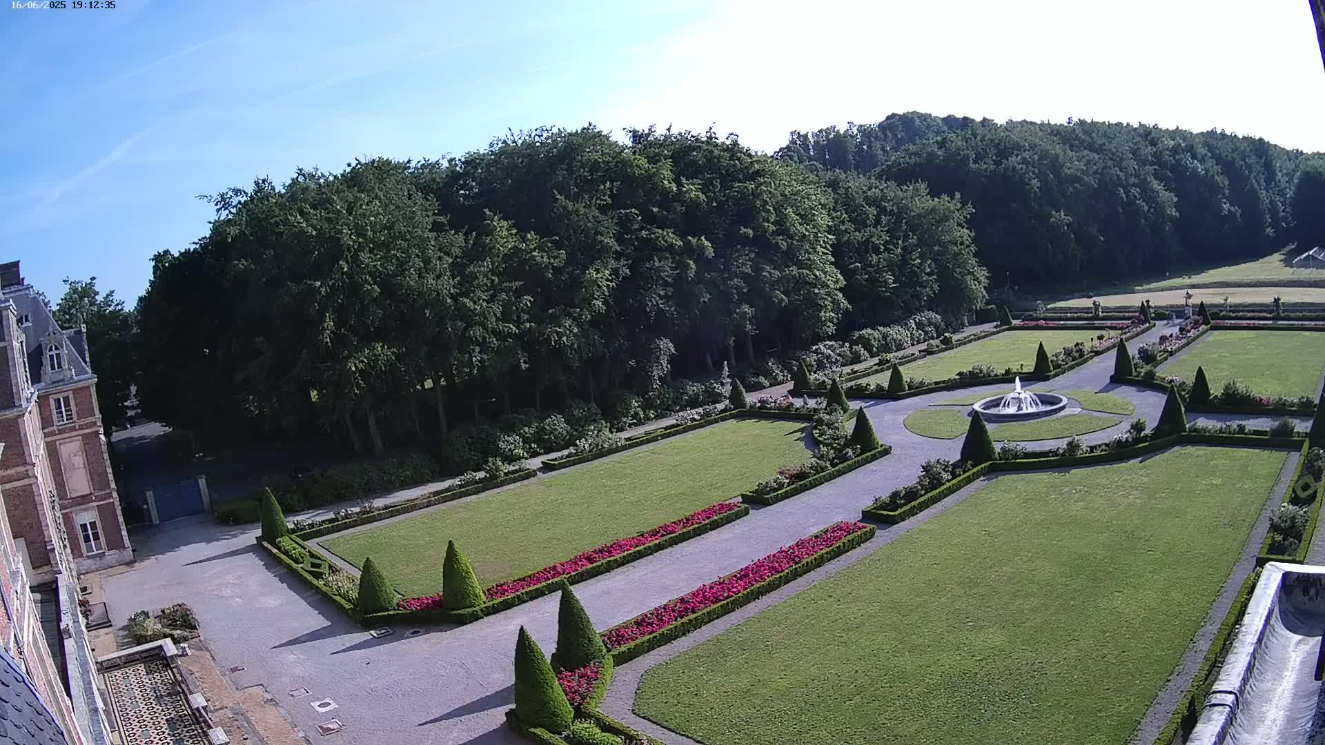 A formal garden with a fountain, manicured lawns, and hedges, is situated beside a large building under a clear blue sky.