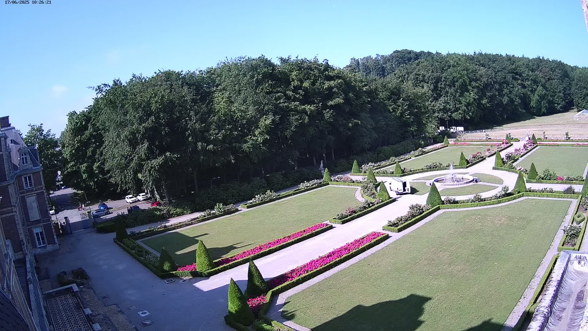 A formal garden with manicured lawns, hedges, and flowerbeds, situated beside a large building and surrounded by a dense forest under a clear, sunny sky.