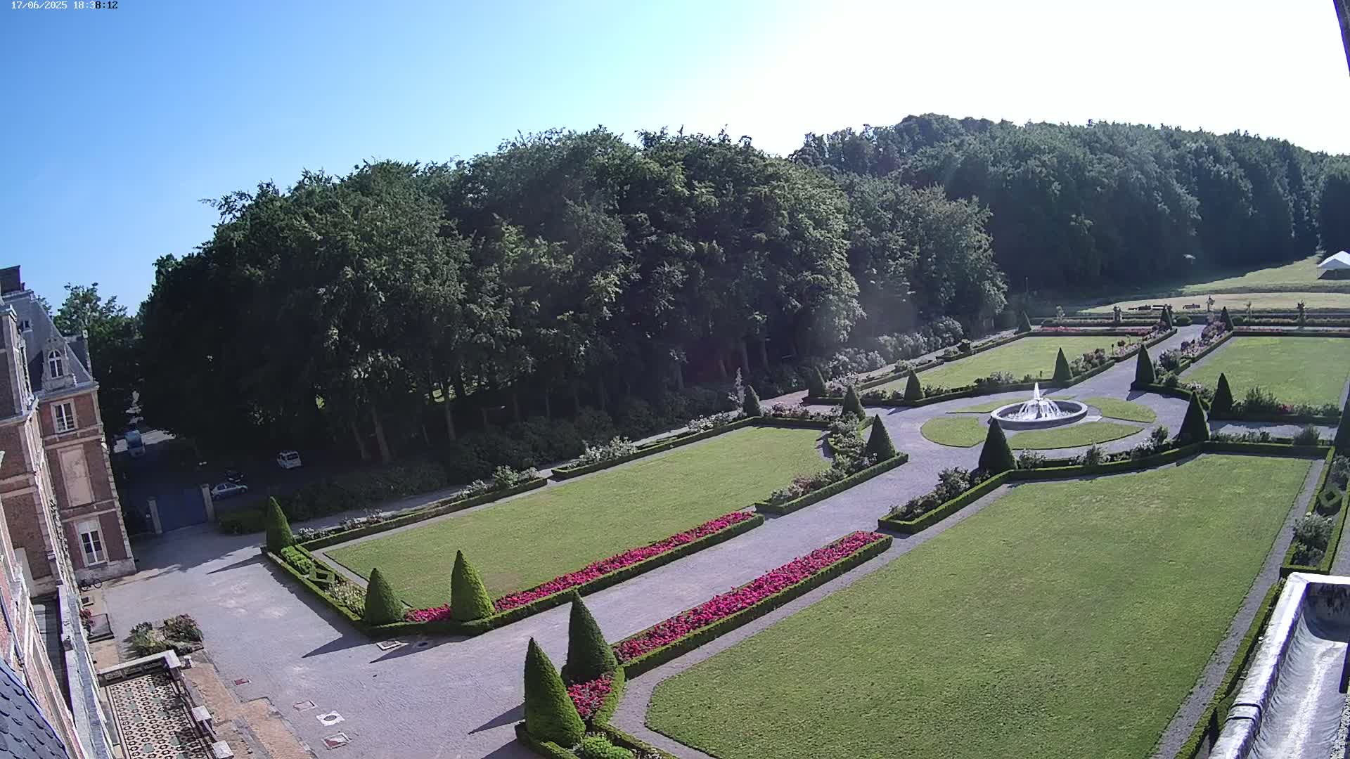 A high-angle view shows a formal garden with a central fountain, manicured lawns, and flowerbeds, adjacent to a large building under a clear, sunny sky.