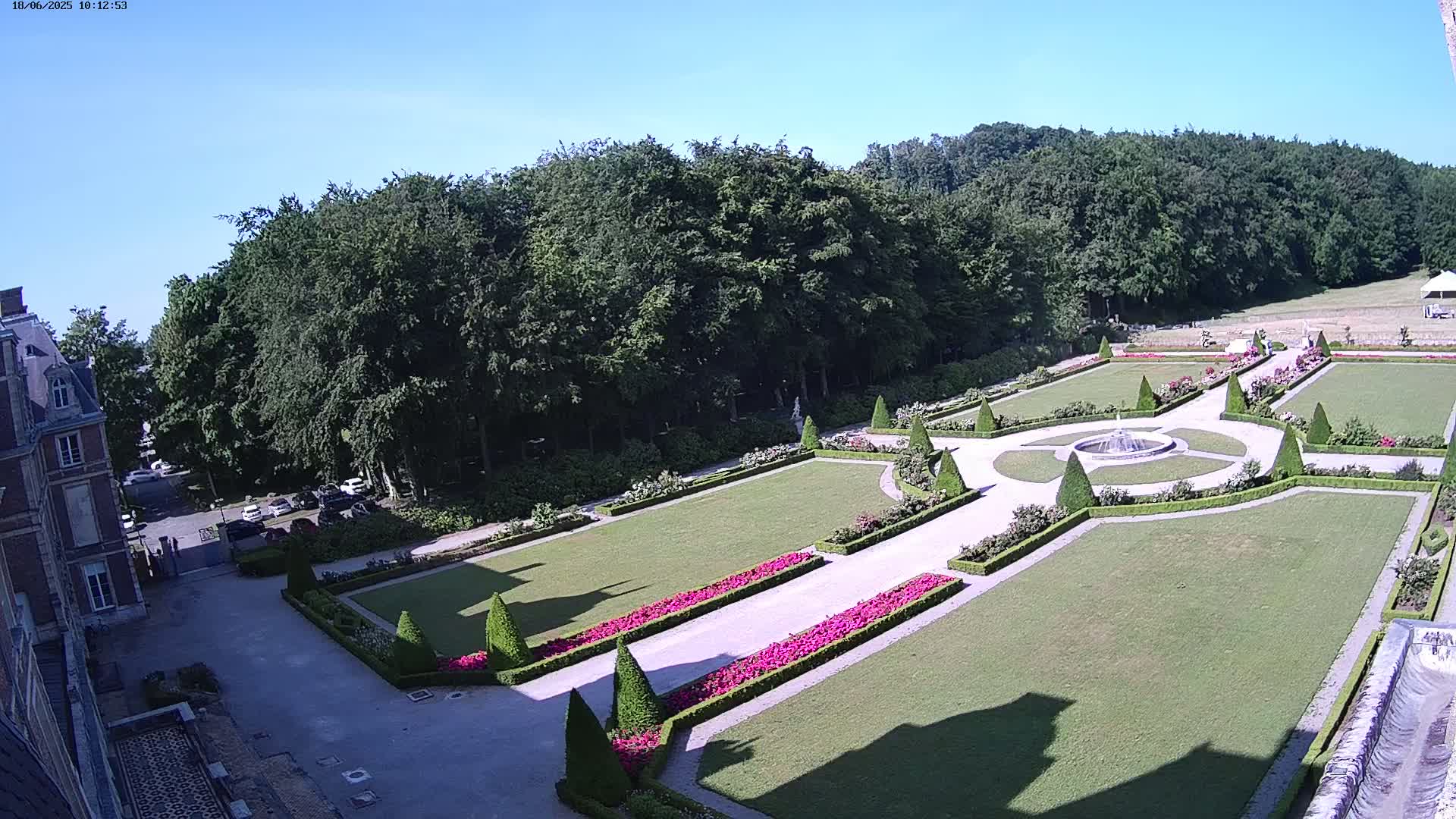 An aerial view shows a formal garden with manicured lawns, hedges, flowerbeds, and a fountain, all set against a backdrop of a dense forest on a sunny day.