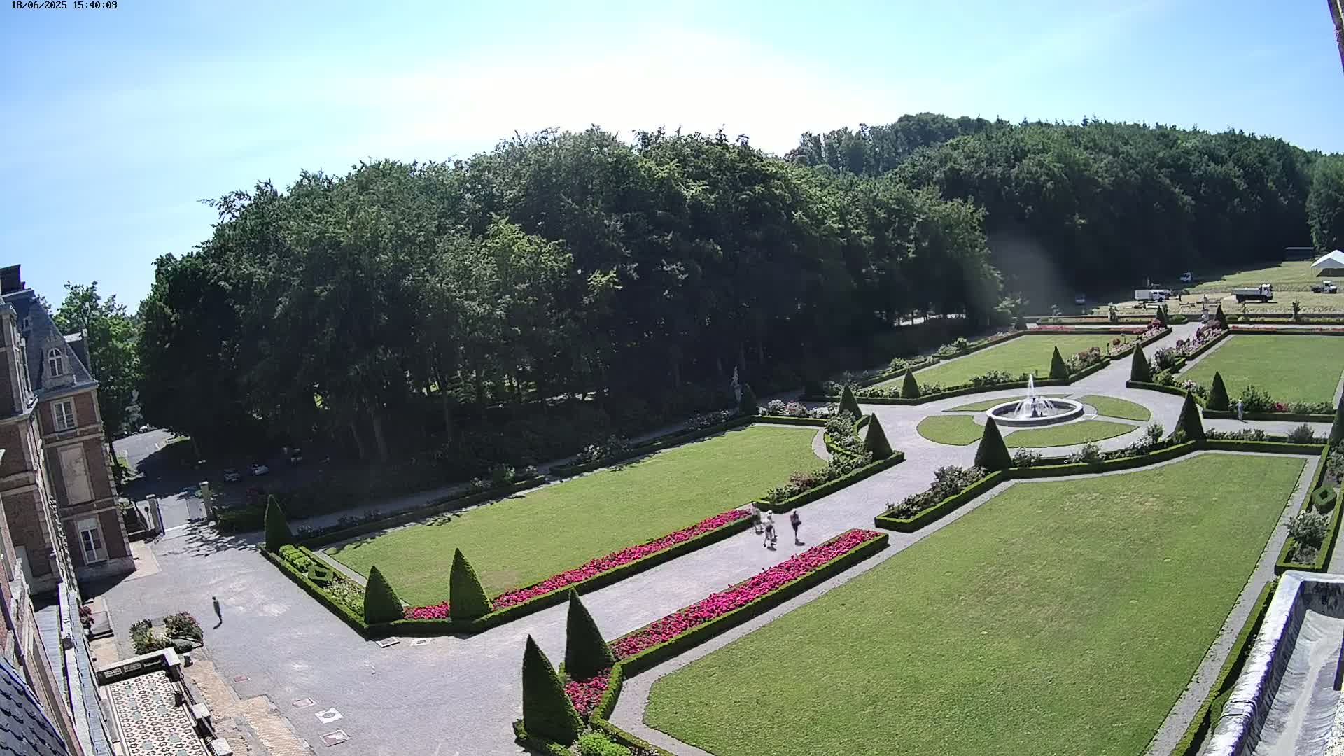 An aerial view shows a formal garden with a central fountain, manicured lawns, and flowerbeds, adjacent to a building, all under a clear, sunny sky.