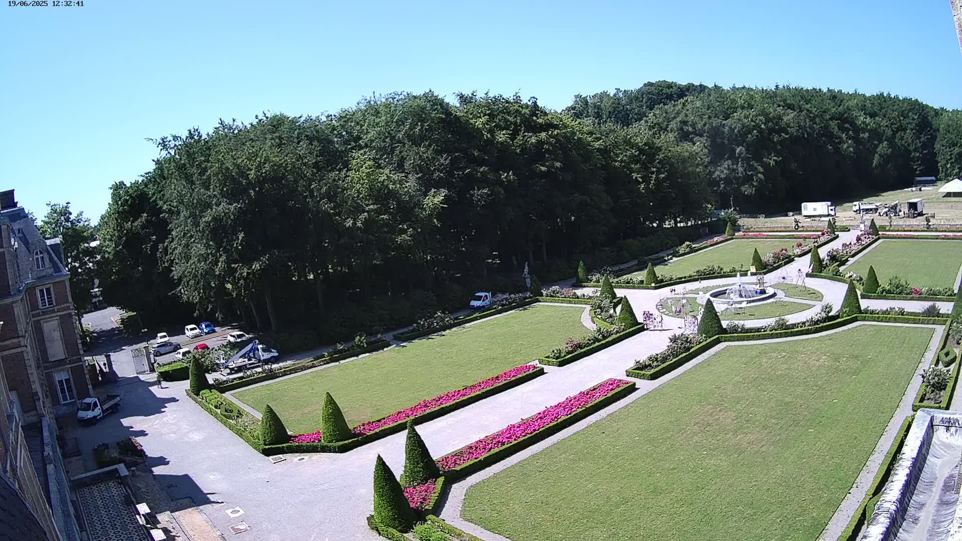 An aerial view shows a formal garden with manicured lawns, flowerbeds, and a fountain, adjacent to a large building and a wooded area under a clear, sunny sky.
