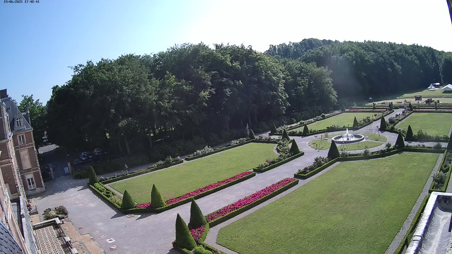 An expansive formal garden with a fountain, manicured lawns, and flowerbeds, situated beside a large building under a clear blue sky.