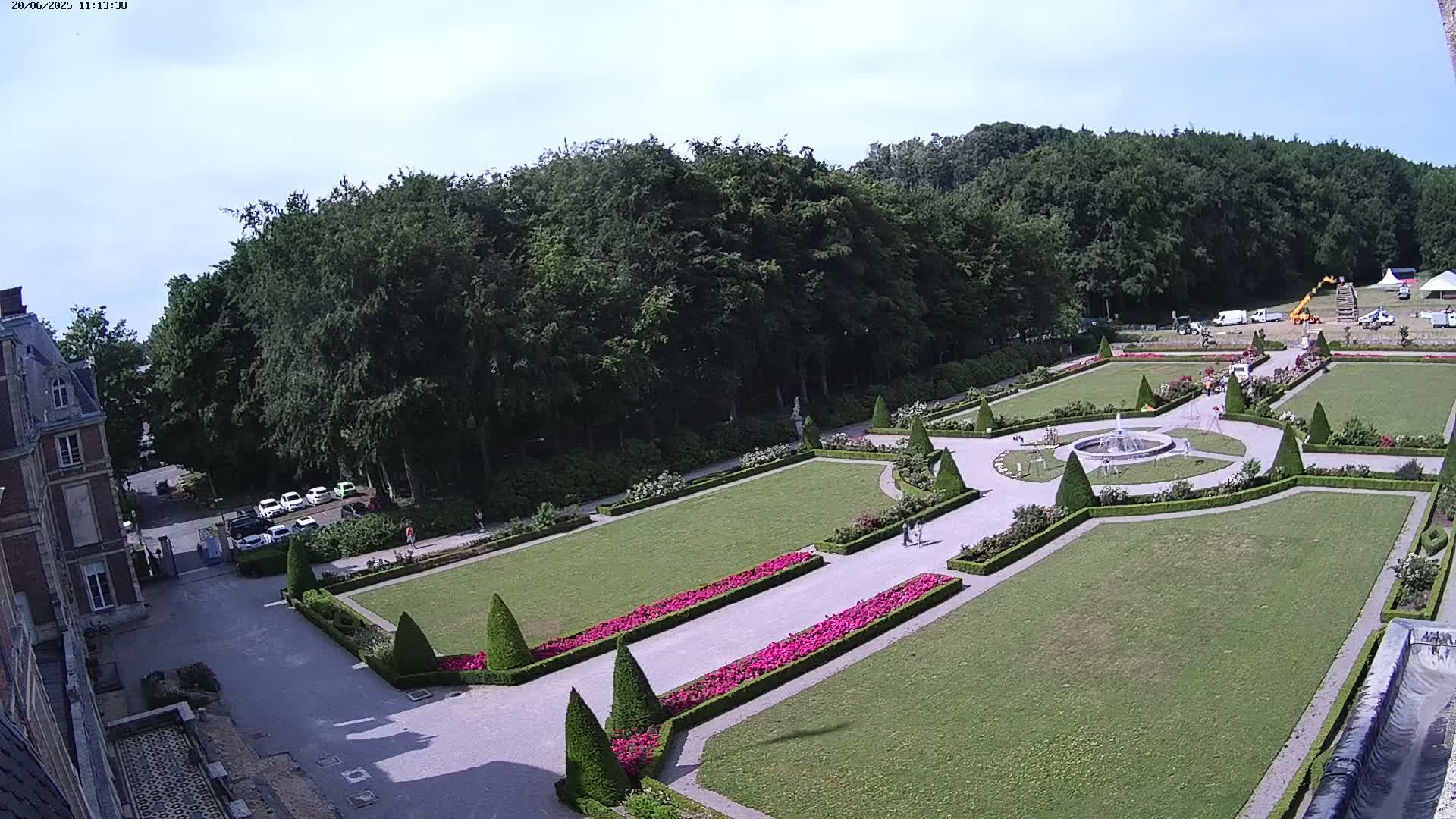 An aerial view shows a formal garden with manicured lawns, hedges, flowerbeds, and a fountain, adjacent to a building and a wooded area under a mostly clear sky.