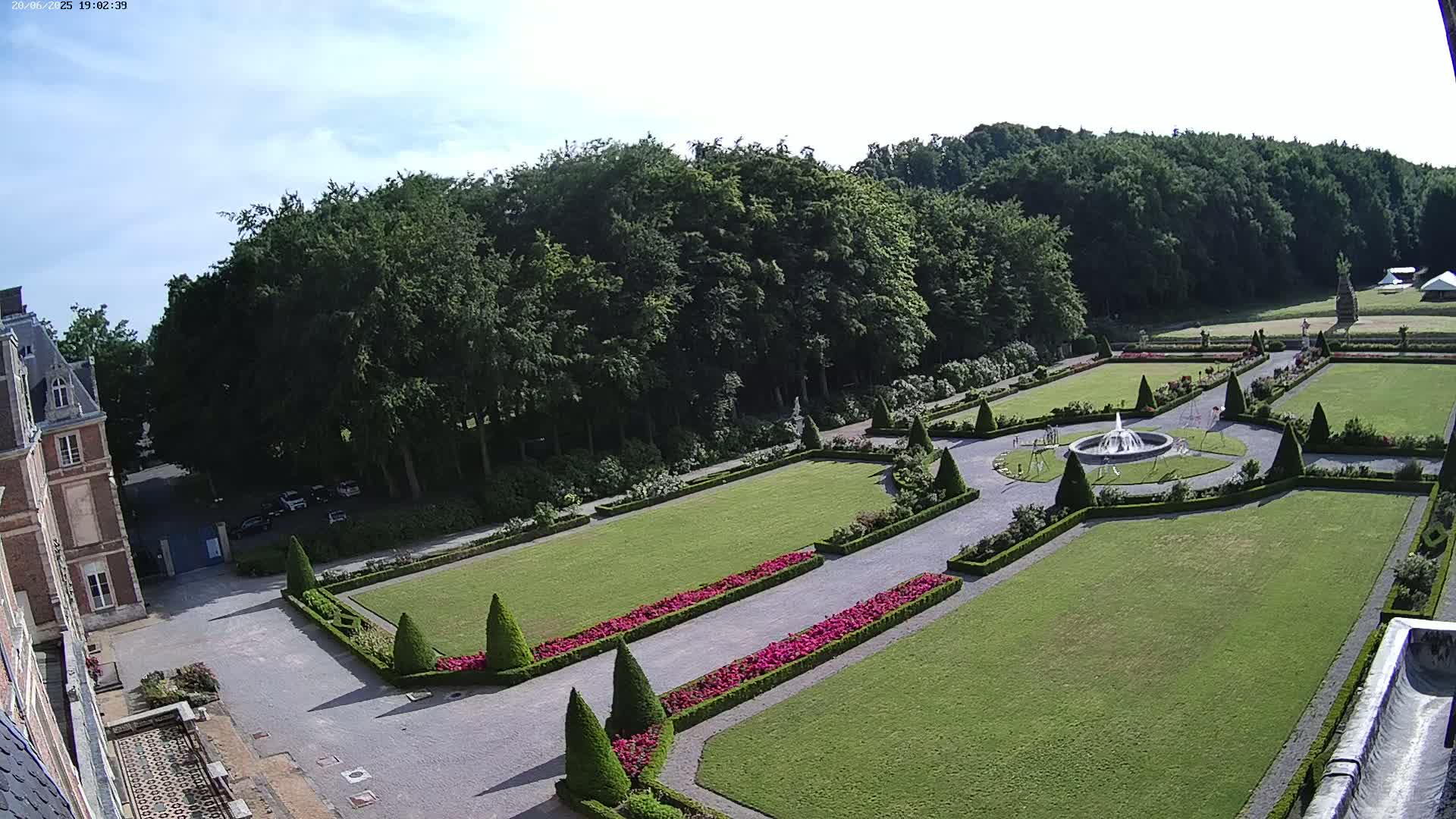 An expansive, formal garden with manicured lawns, flowerbeds, and a central fountain is situated next to a large brick building under a mostly clear sky.
