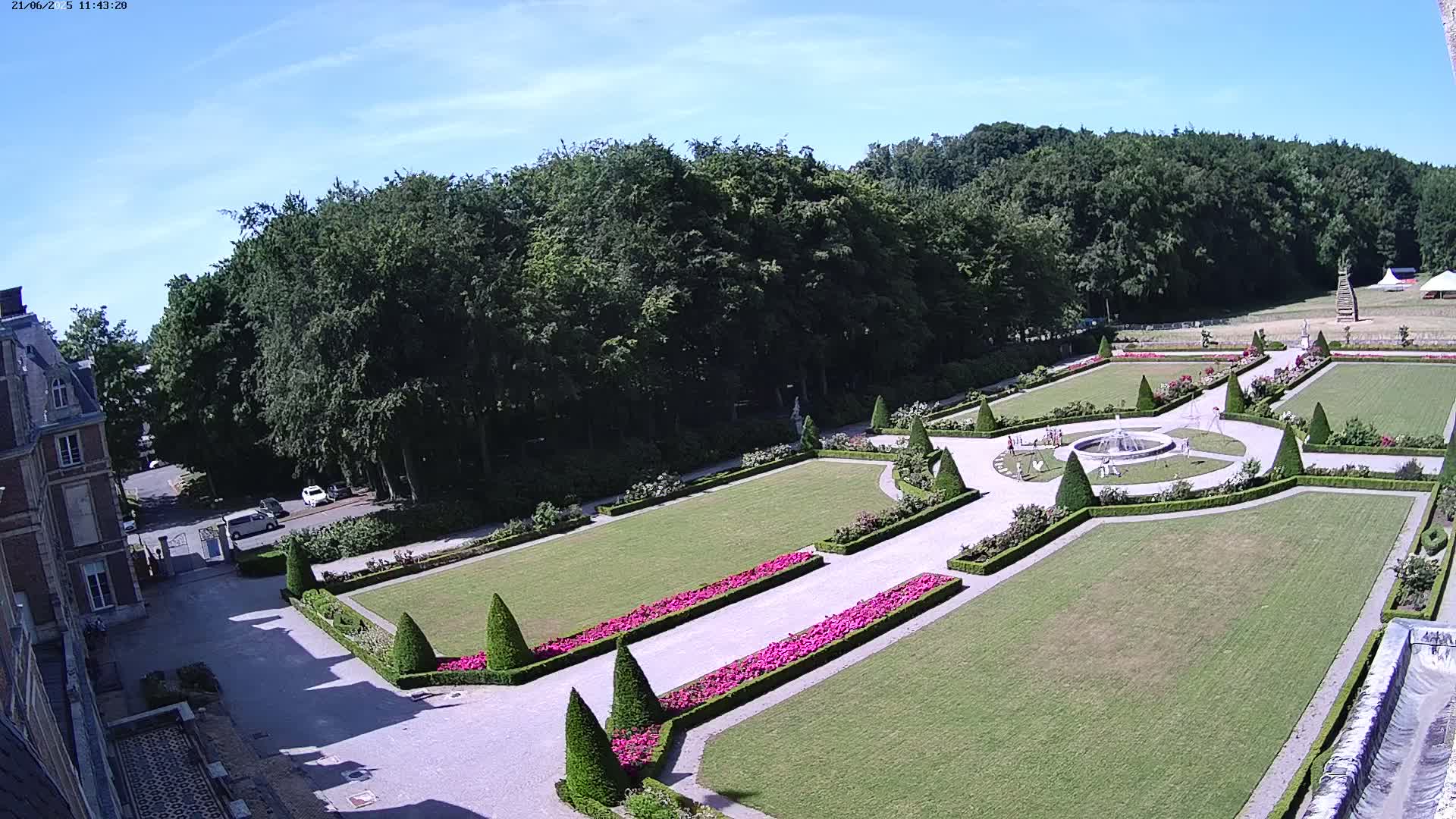 An aerial view shows a formal garden with manicured lawns, flowerbeds, and a fountain, situated beside a large building under a clear blue sky.