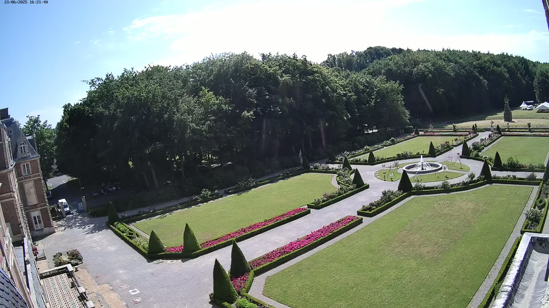 A formal garden with a central fountain, manicured lawns, and hedges, is adjacent to a building under a mostly sunny sky.