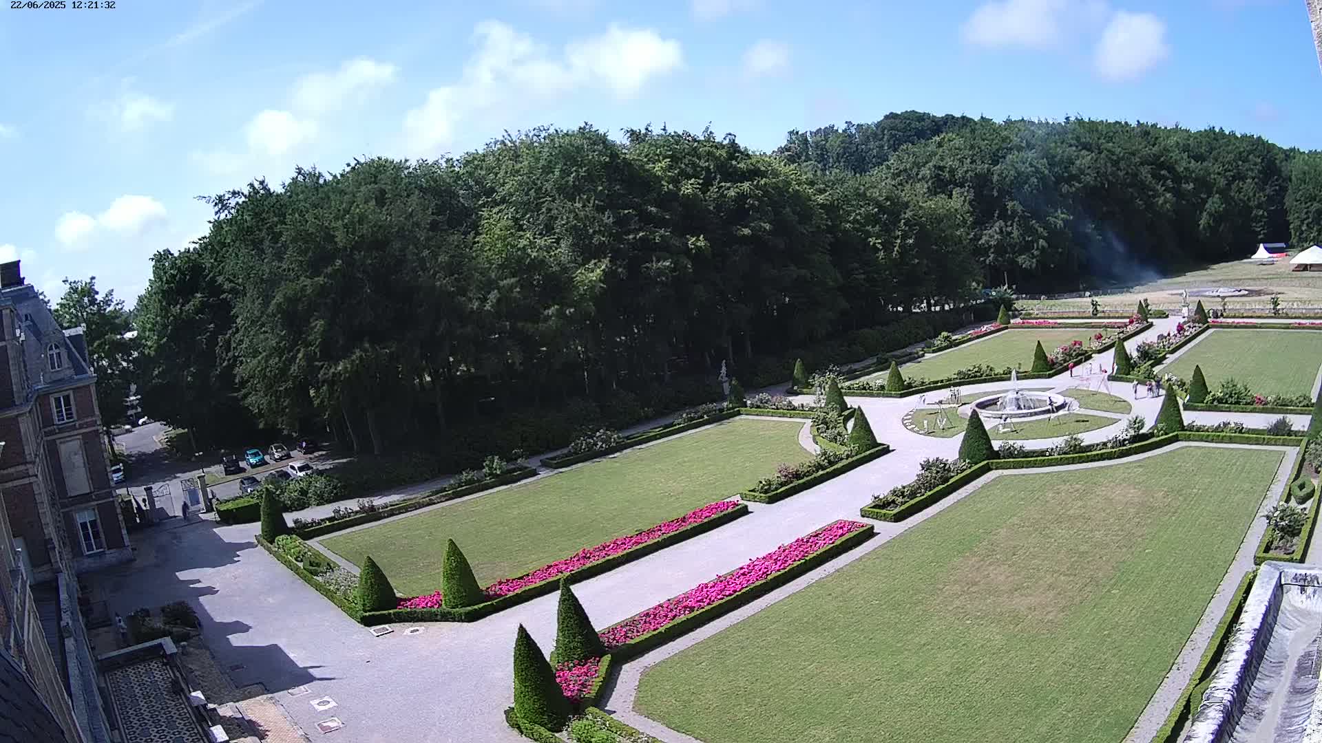 An aerial view shows a formal garden with manicured lawns, hedges, flowerbeds, and a fountain, situated next to a large building under a mostly sunny sky.