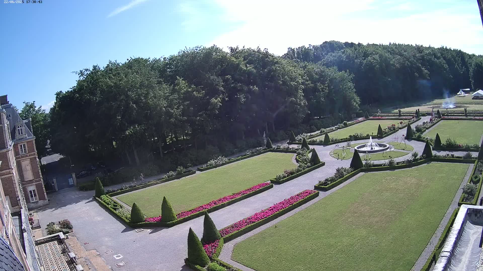An elevated view of a formal garden with a fountain, manicured lawns, and flower beds, adjacent to a chateau under a clear blue sky.