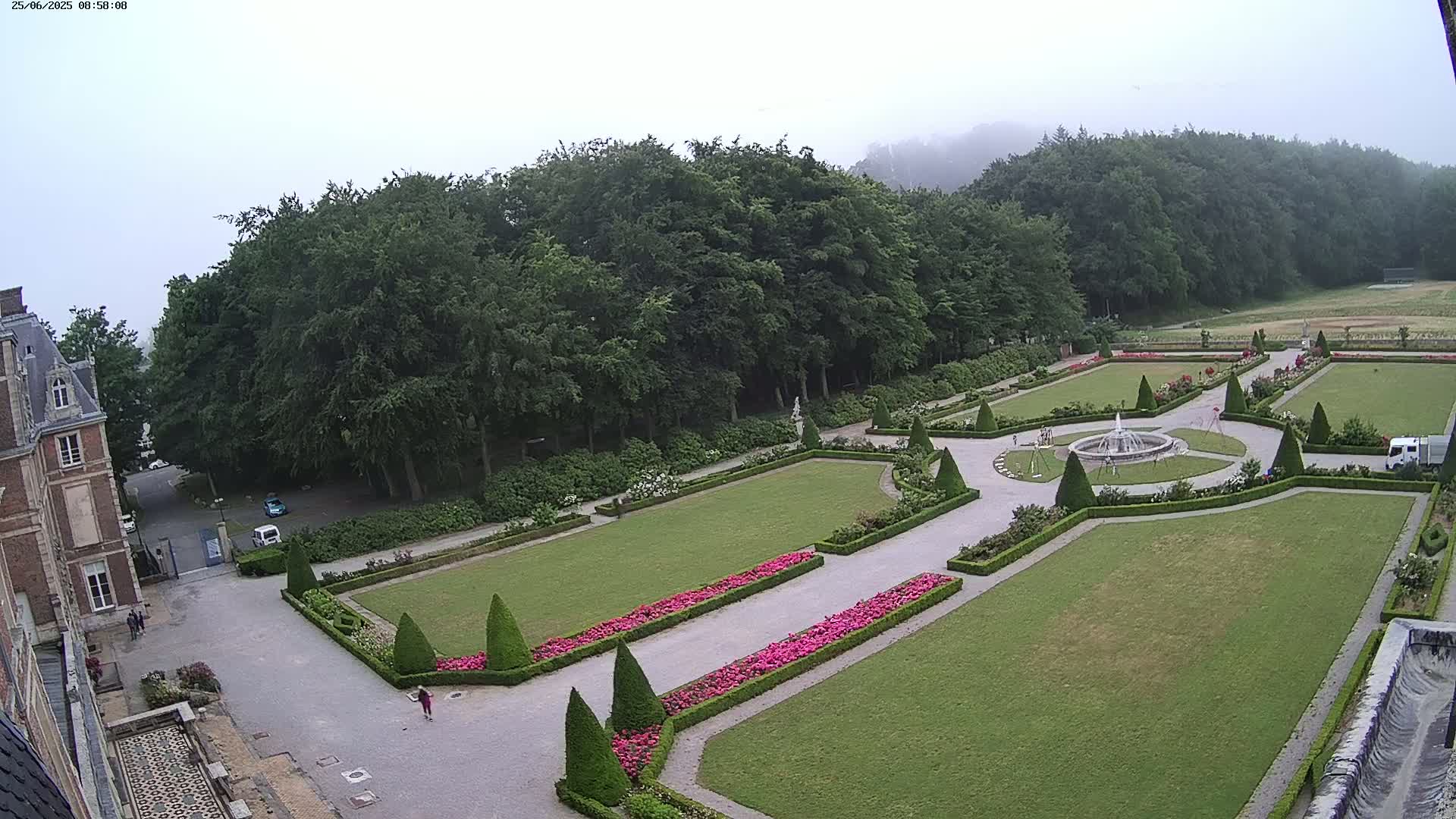 An expansive formal garden with a central fountain is situated next to a large building, all under a hazy sky.