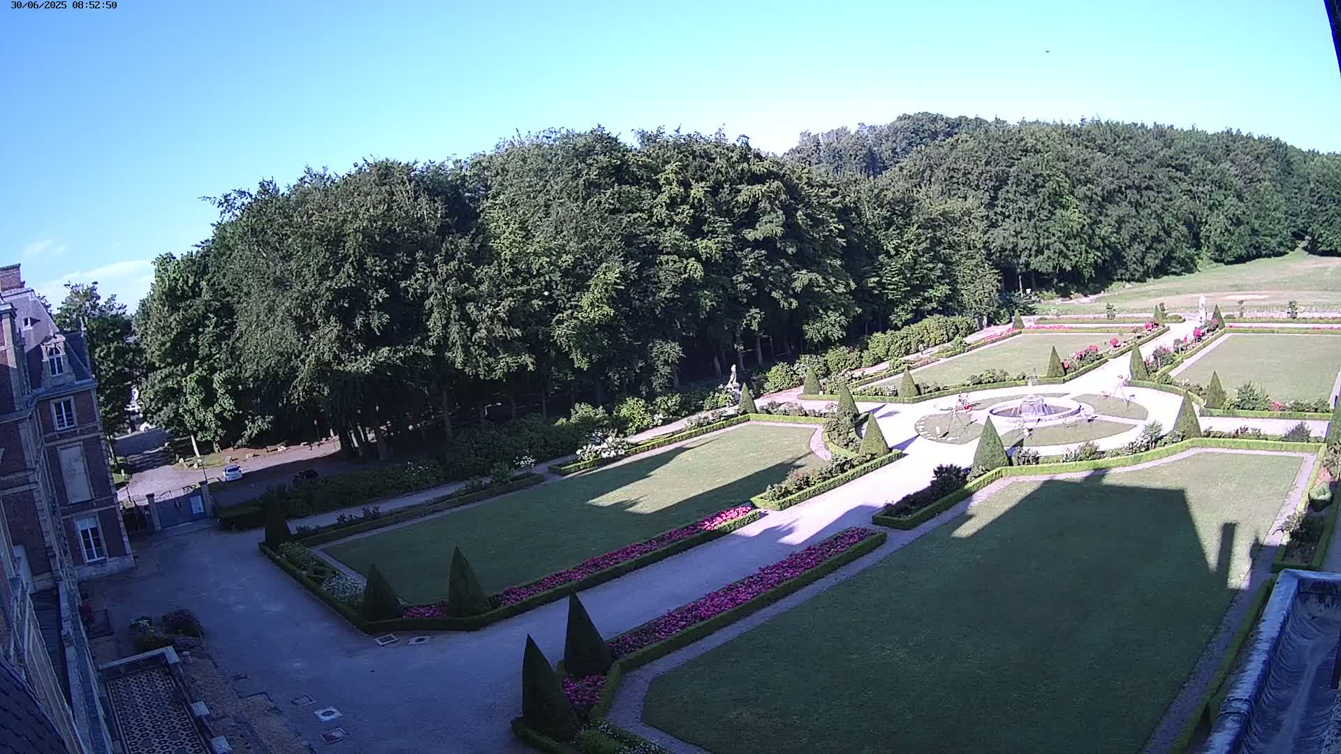 An aerial view shows a formal garden with manicured lawns, hedges, flowerbeds, and a fountain, bordered by a large forest under a clear, sunny sky.