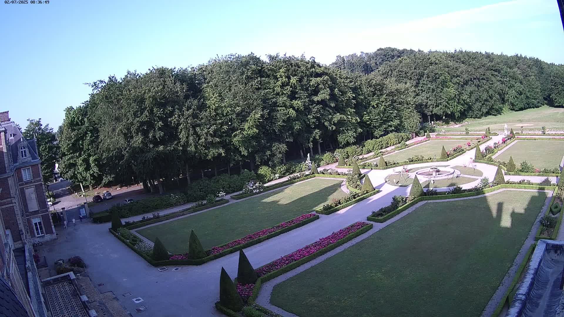 An expansive formal garden with manicured lawns, flowerbeds, and hedges is situated next to a large building under a clear, sunny sky.