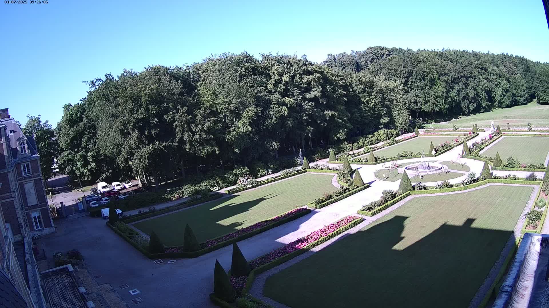 An aerial view shows a formal garden with manicured lawns, hedges, and a fountain, situated next to a large building under a clear blue sky.