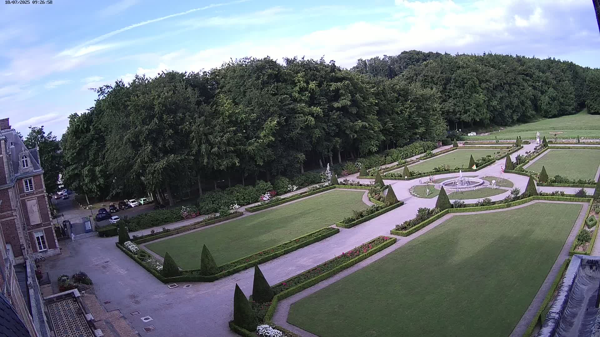 An aerial view shows a formal garden with geometrically-shaped lawns, hedges, and a fountain, set against a backdrop of a forest under a partly cloudy sky.