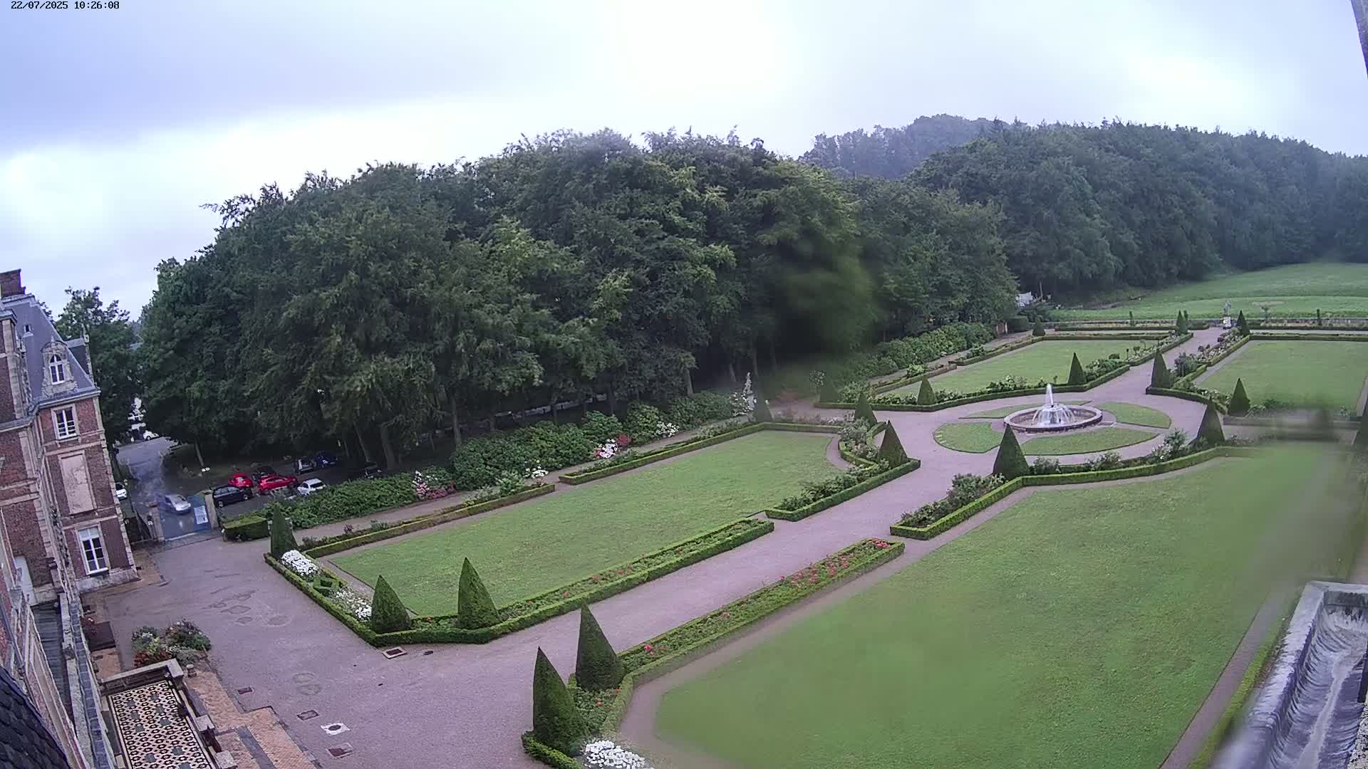 An overcast day reveals a formal garden with a central fountain, manicured lawns, and hedges, adjacent to a large building.