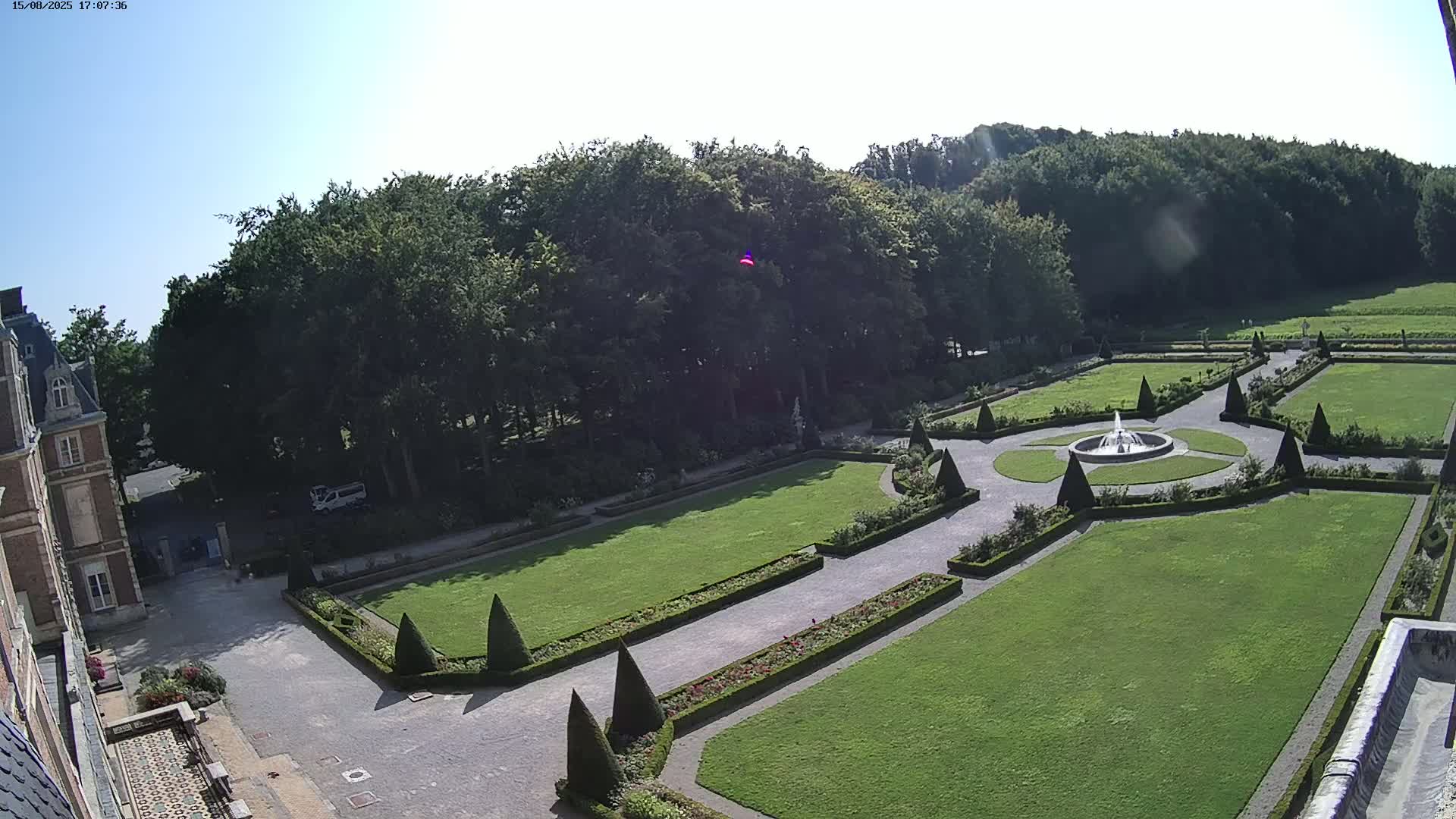 An overcast day reveals a formal garden with a central fountain, manicured lawns, and hedges, adjacent to a large building.