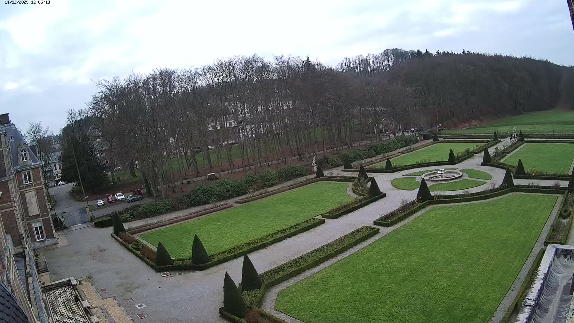 An overcast day reveals a formal garden with a central fountain, manicured lawns, and hedges, adjacent to a large building.